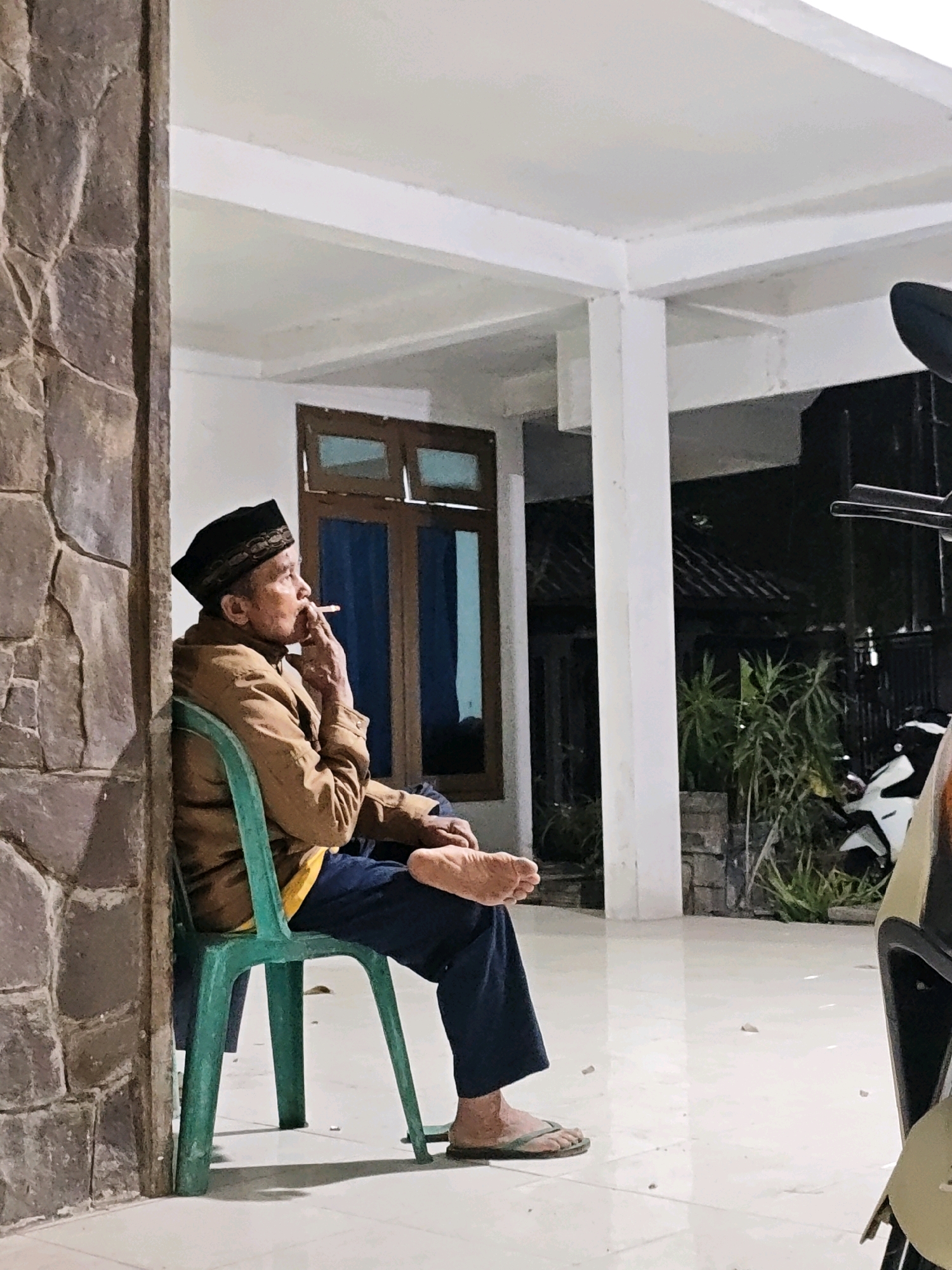 An elderly man, wearing a black peci hat and a brown jacket, sits on a green plastic chair on a tiled patio in Gurah, Indonesia. He is smoking a cigarette, holding it with his right hand and bringing it to his mouth. The man is leaning against a stone wall on his left, and his legs are crossed, with his right foot bare and wearing a flip-flop. Behind him, a window with wooden frames and blue panes is visible. The patio is covered by a white, modern-style overhang supported by white pillars. In the background, a motorcycle is parked, and vegetation can be seen. The scene appears to be an outdoor area of a residential or public building.