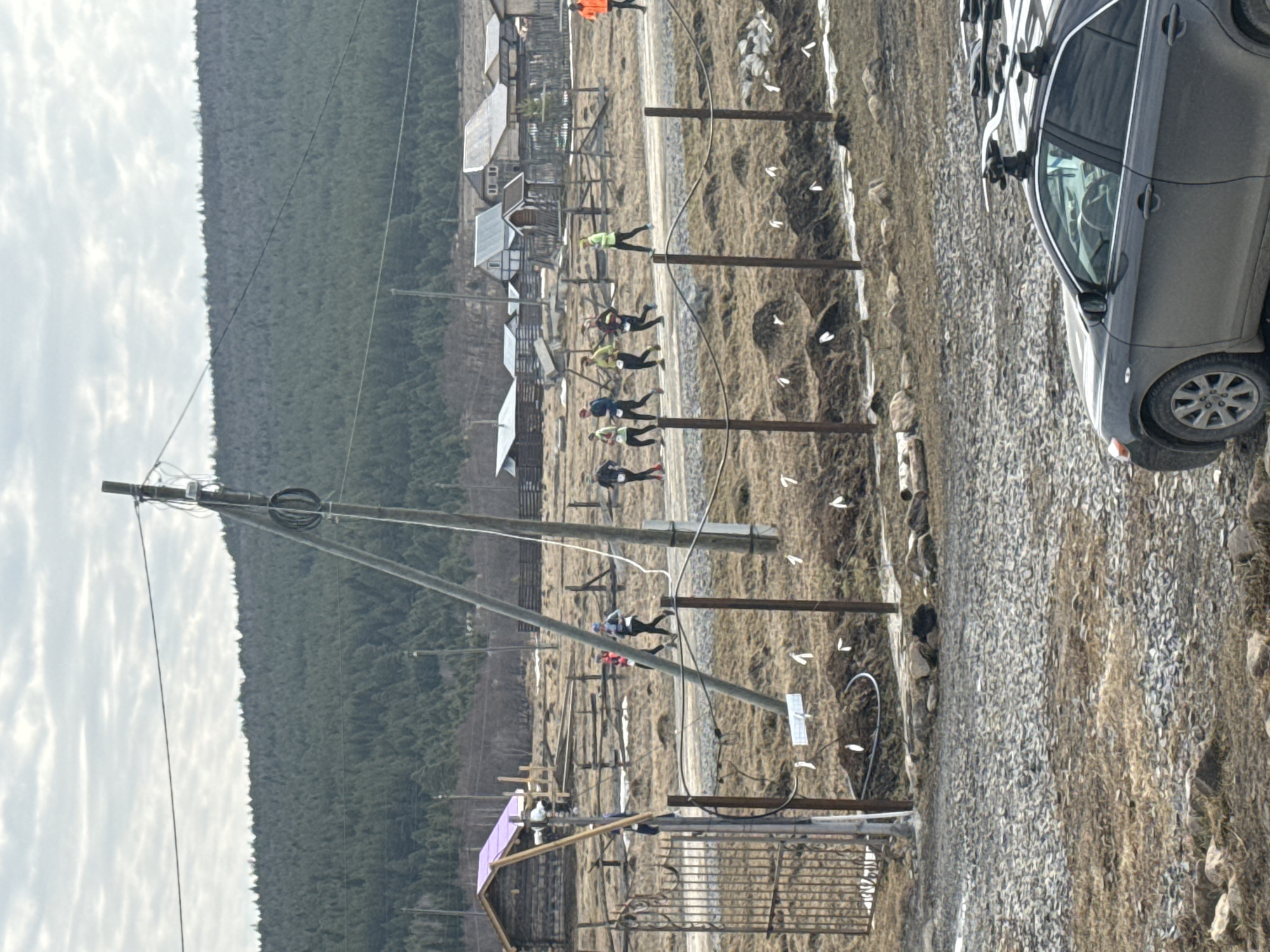 A landscape scene near the village of Tyulyuk in Russia shows a gravel road with a gray car parked beside it on the right. The car has roof racks. To the left of the road is a series of wooden posts, likely part of a fence, and a group of people are running beside the posts. These people seem to be athletes participating in some sort of race or activity. Behind them is a small village with wooden buildings, and in the background, a dense forest stretches along a hillside. An electric pole with wires is visible on the left side of the image. The sky is partly cloudy, suggesting an overcast day.
