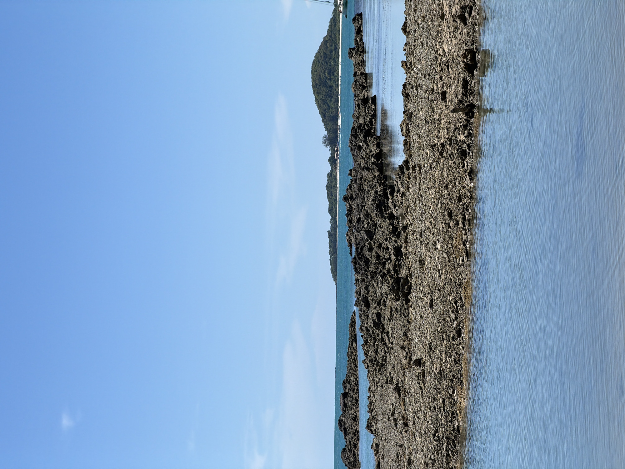 The image depicts a serene coastal scene in Baan Thong Krut, Thailand. In the foreground, shallow, calm blue water reflects the bright sky. This is followed by a rocky, uneven shoreline composed of dark, coarse stones and pebbles, with some patches of sand visible. Further out, the turquoise ocean stretches towards the horizon, meeting a clear blue sky with a few wispy clouds. In the distance, a lush green, tree-covered island with a white sandy beach is visible, dotted with small buildings and boats. The overall environment is natural and undeveloped, suggesting a tranquil, tropical setting. There are no people or animals visible in the image.