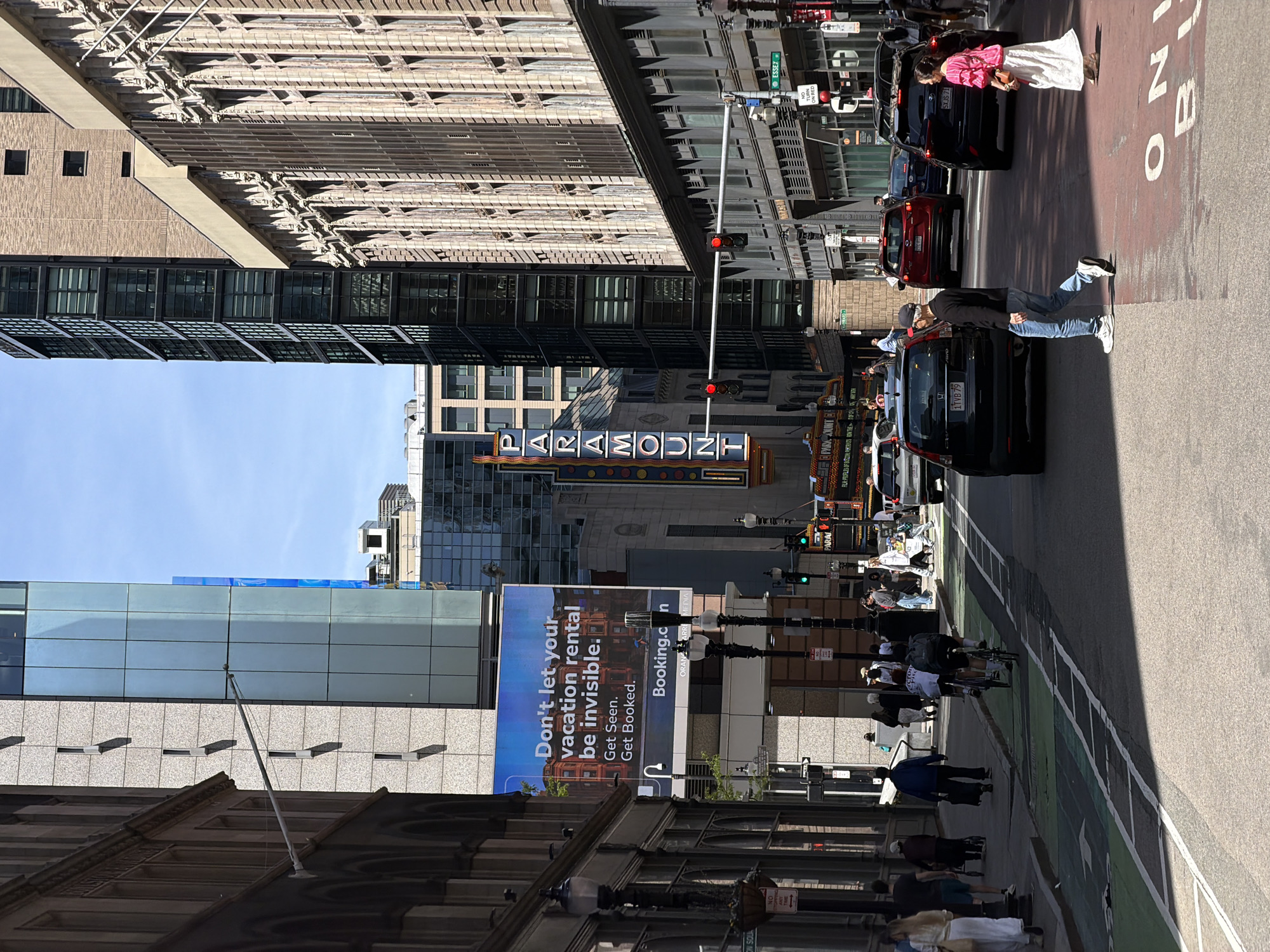 The image shows a street scene in Boston, USA, featuring the Paramount Theater's sign and surrounding city infrastructure. The theater sign is prominently displayed, and adjacent buildings include modern architecture and older brick structures. A large advertisement from Booking.com is visible on one of the buildings.
The street is active with pedestrian and vehicular traffic. People are walking on the sidewalks, and several cars are moving along the road. Traffic lights are present, indicating the controlled flow of vehicles. The layout of the street includes dedicated bicycle lanes.