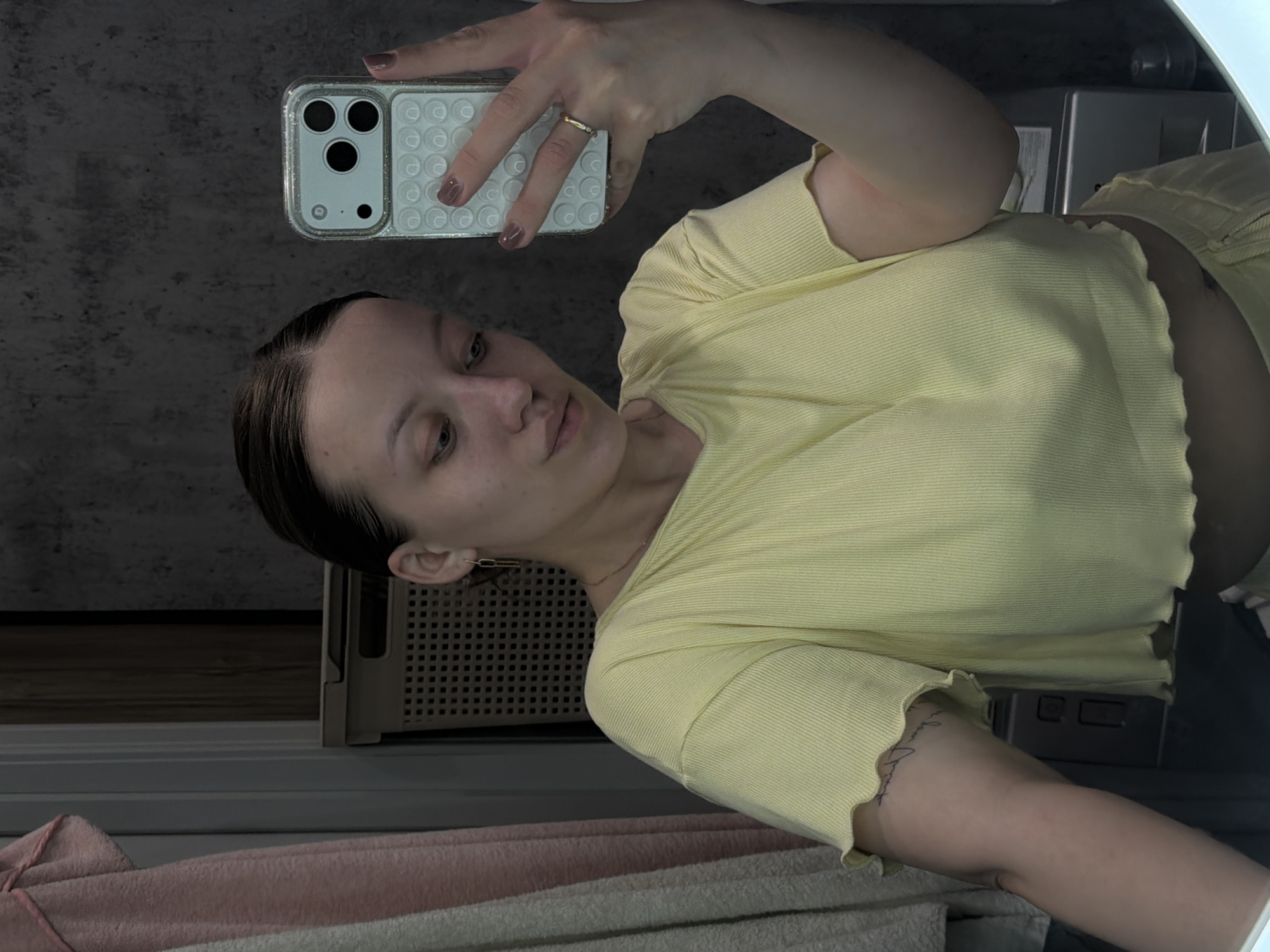 A woman is taking a selfie in a bathroom. She is wearing a yellow crop top. She is holding a phone with a patterned case in her right hand. Her left arm is visible, showing a tattoo. The bathroom appears to be modern with a grey countertop and a laundry hamper visible in the reflection. The location of the image is not discernible to be the city of Reshetnikovo, Russia from the contents.
