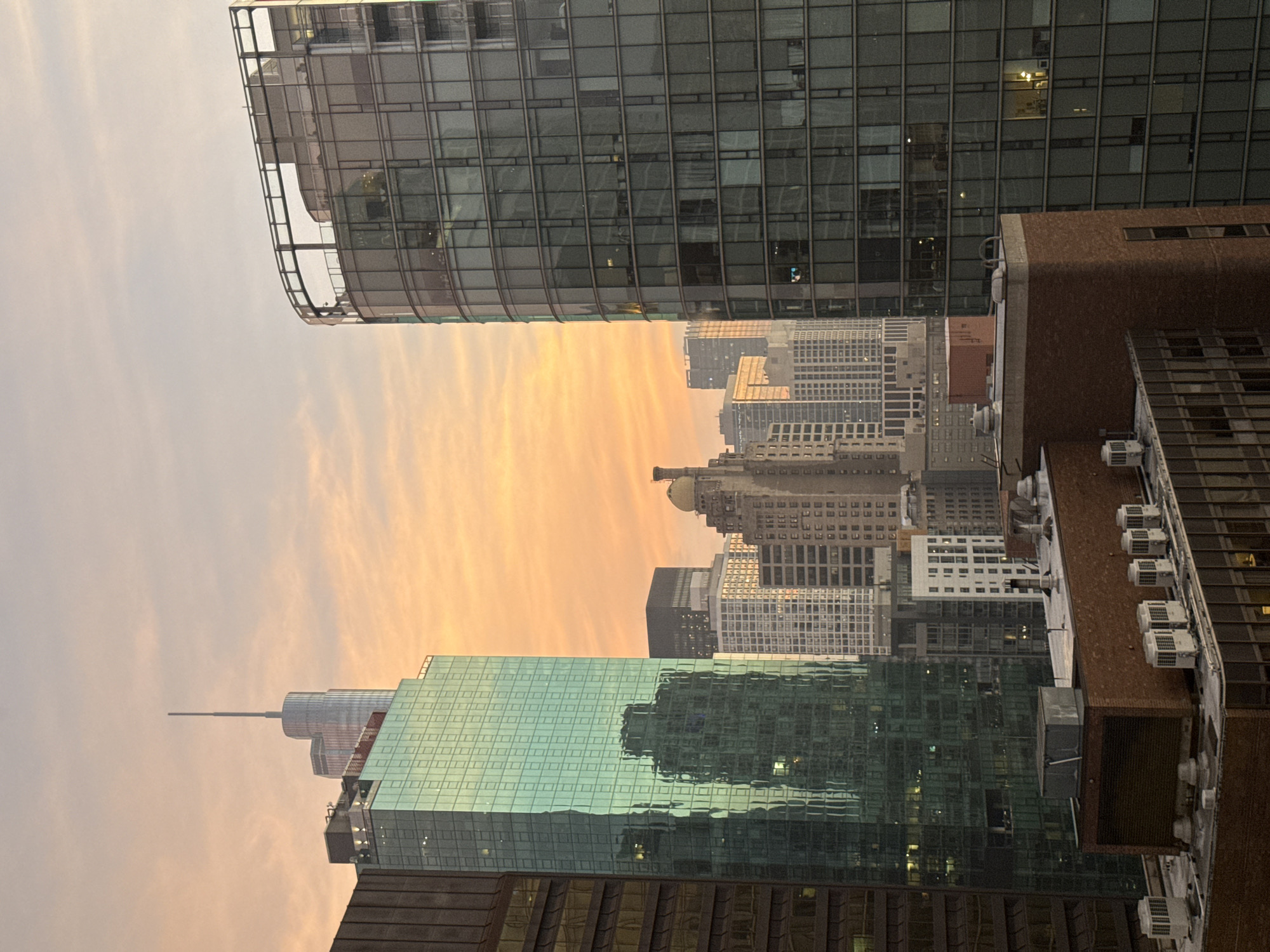 This is a photograph of a city skyline at sunset, likely in Chicago, Illinois, United States of America, given the architectural style of the buildings. The sky is filled with warm, orange and pink hues, indicative of a sunset, with wispy clouds present. In the foreground and mid-ground, various skyscrapers are visible.
On the left, a tall, rectangular building with a teal-colored glass facade dominates the view. Its reflective surface mirrors other buildings. Adjacent to it and slightly behind is a building with a darker facade. In the center, several buildings of varying heights and architectural styles are clustered together. One particularly notable building in this cluster has a distinctive domed roof and older, stone-like facade. To the right, a large, curved skyscraper with a dark glass exterior stands prominently. Its windows reflect the sky and surrounding structures.
In the foreground, the rooftops of lower buildings are visible. These include brick structures with various mechanical units, such as air conditioning units, and what appear to be ventilation pipes. The overall scene depicts an urban environment during the transition from day to night, with artificial lights beginning to appear in some of the building windows. No people are discernible in the image.