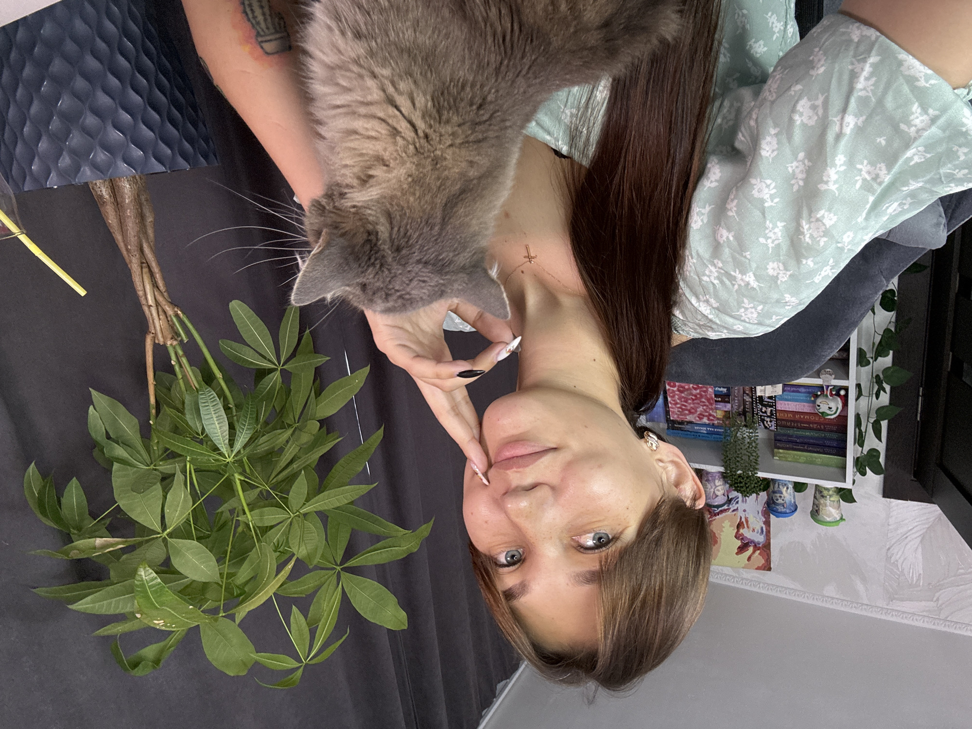 A young woman with light brown hair pulled back in a ponytail and blue eyes is seated in a chair, looking at the camera. She is wearing a light green floral print blouse. A grey cat is perched on her lap, facing away from the camera. In the background, a bookshelf filled with books and figurines is visible, along with a potted plant and a dark curtain. The scene appears to be indoors, likely in a home setting. The presence of books and decorative items suggests a personal living space. The lighting is soft, and the overall atmosphere is relaxed.