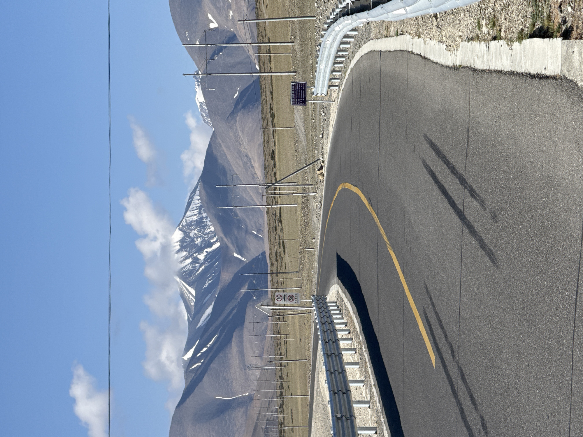 The image captures a sweeping view of a high-altitude mountain road in Gyirong County, China, under a bright, clear blue sky with scattered white clouds. A paved asphalt road, featuring a prominent yellow dividing line, curves gently to the right in the foreground. Distinct dark shadows, possibly cast by unseen overhead objects or vehicles, stretch across the road surface. A silver metal guardrail runs along the outer edge of the curve, bordering rocky and barren terrain.
In the mid-ground and background, majestic, snow-capped mountains dominate the horizon, their rugged peaks showcasing patches of white snow. The lower slopes of these mountains appear dry and sparsely vegetated with brownish scrub. A series of tall, slender poles, likely for power lines or communication, line the left side of the road, extending into the distance. A thin black wire is visible horizontally across the upper portion of the sky.
Two signs are visible on the left side of the road. One is a blue rectangular sign with white Chinese characters, which appears to be an informational or directional sign. Adjacent to it is a circular white sign with a red border, displaying the numbers "80" and "60", indicating speed limits. The bright sunlight and sharp shadows suggest the time of day is likely midday or early afternoon. The scene conveys a sense of vastness and remote natural beauty characteristic of a mountainous region.