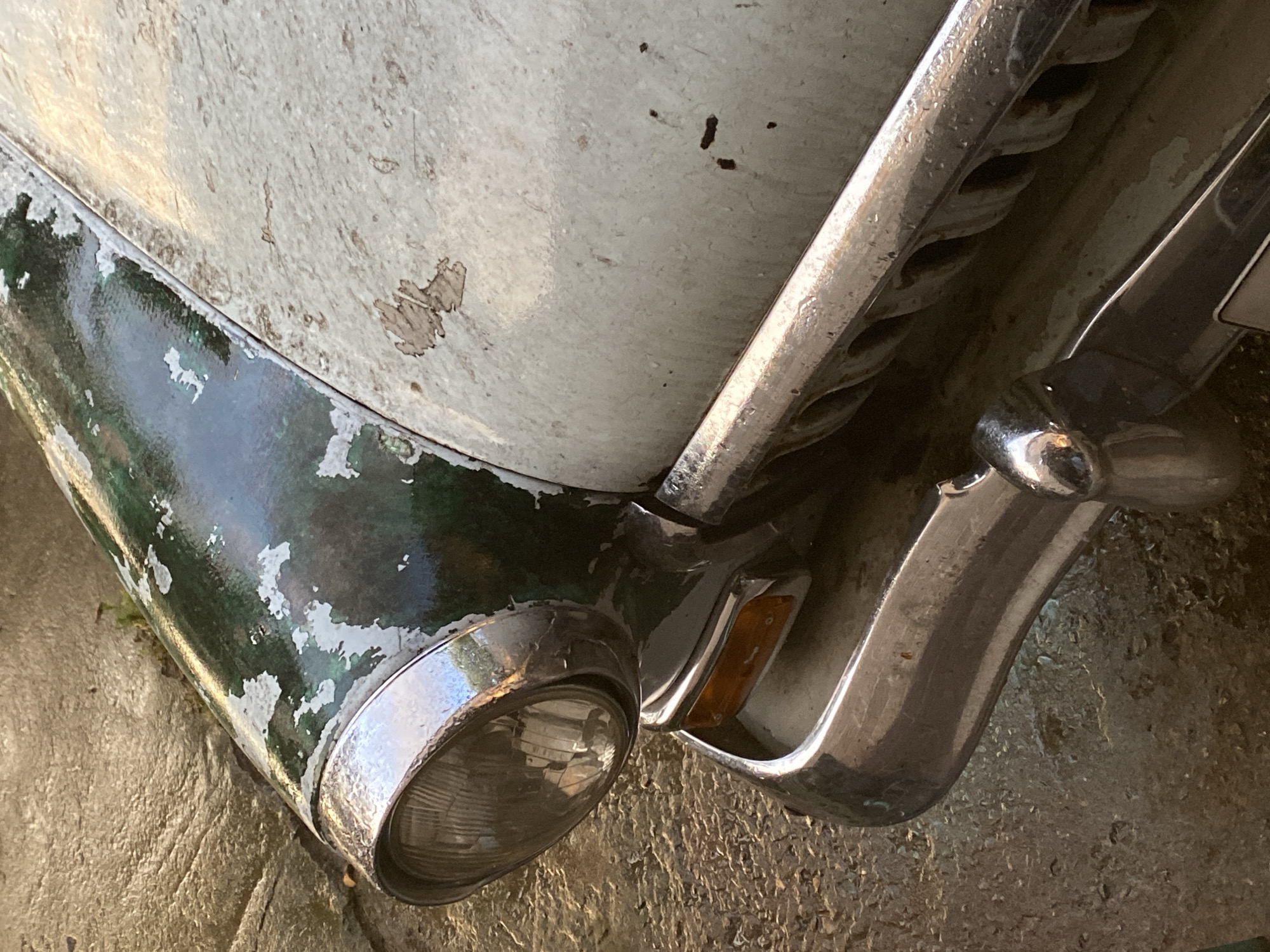 The image shows a close-up view of the front of an old, weathered car, likely parked on a cobblestone or rough concrete surface. The car's paint is peeling, revealing layers of green and white underneath. A chrome bumper and headlight assembly are visible, showing signs of age and wear. The background is blurred and suggests an outdoor environment, possibly a street or yard. There are no people or visible activity. The location is Dinskaya, Russia.