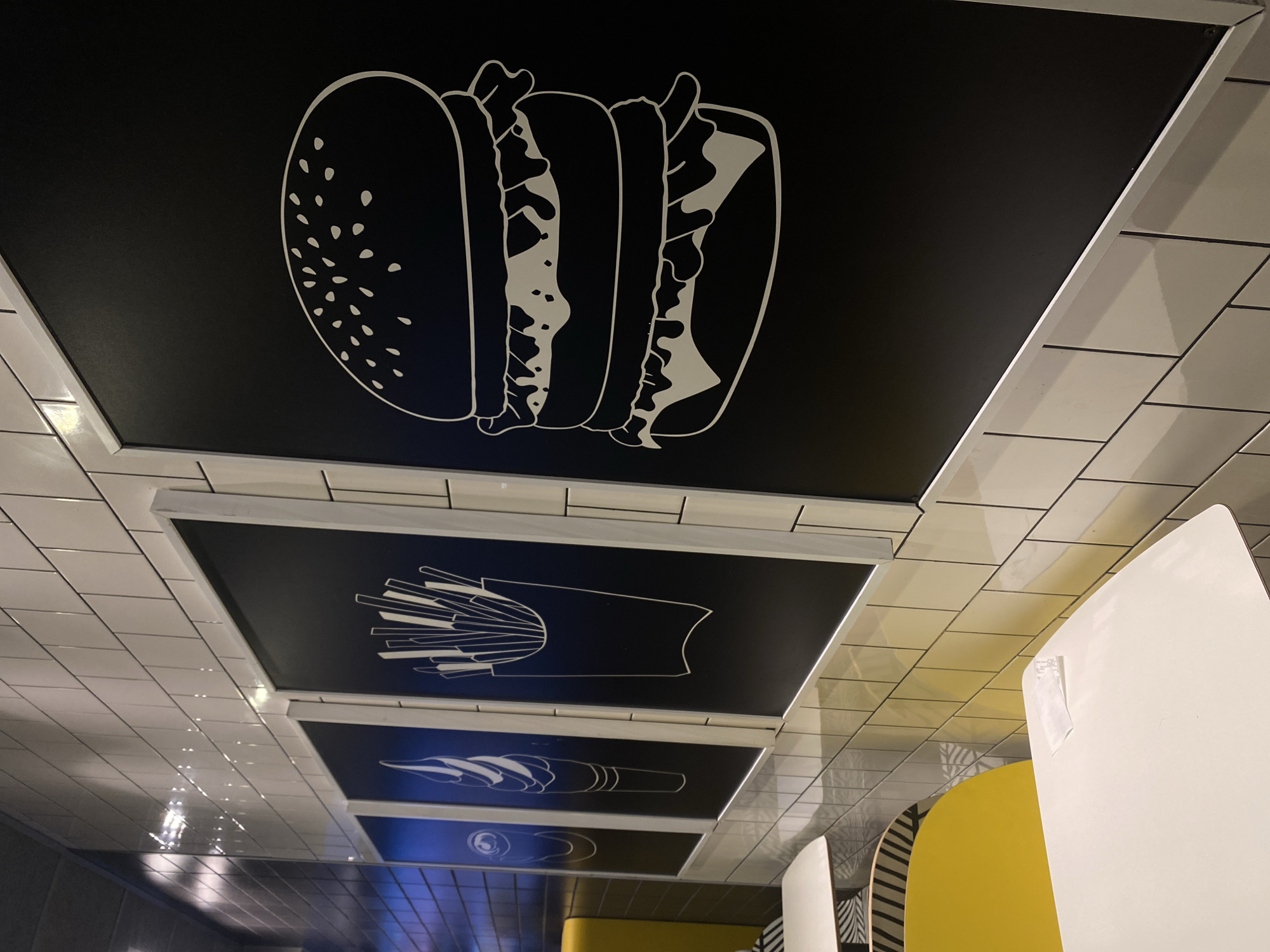 The image shows a side view of a restaurant interior in Balashikha, Russia. The walls are tiled with white subway tiles. Mounted on the wall are four framed black boards with white line drawings of food items: a can, an ice cream cone, French fries in a carton, and a large hamburger. In the foreground, a white table with a yellow chair is visible. The overall impression is of a clean and modern fast-food establishment.