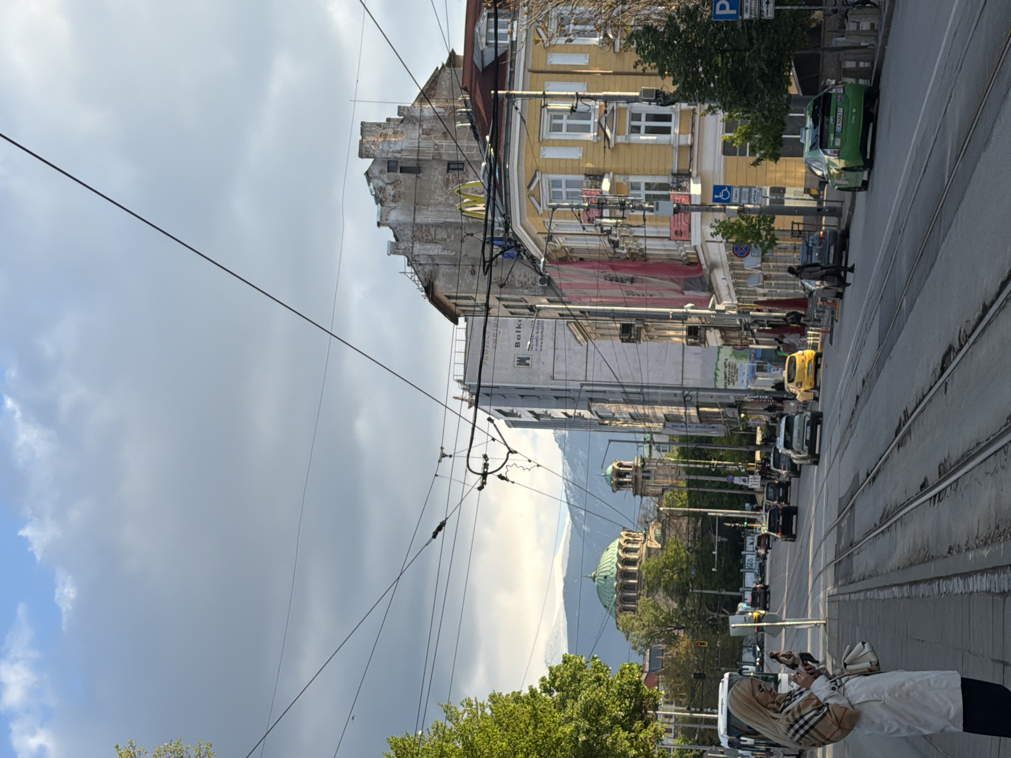 The image shows a city street scene in an unknown location. The street has tram tracks and is lined with buildings of various heights and architectural styles, including some with yellow facades. A McDonald's sign is visible on one of the older buildings. Cars and pedestrians are present on the street. Overhead wires are visible, possibly for trams or other electric transport. In the distance, a building with a green dome is visible, suggesting it could be a religious or governmental building. The sky is cloudy with a mix of blue and grey, indicating overcast weather. A woman is seen holding a phone.