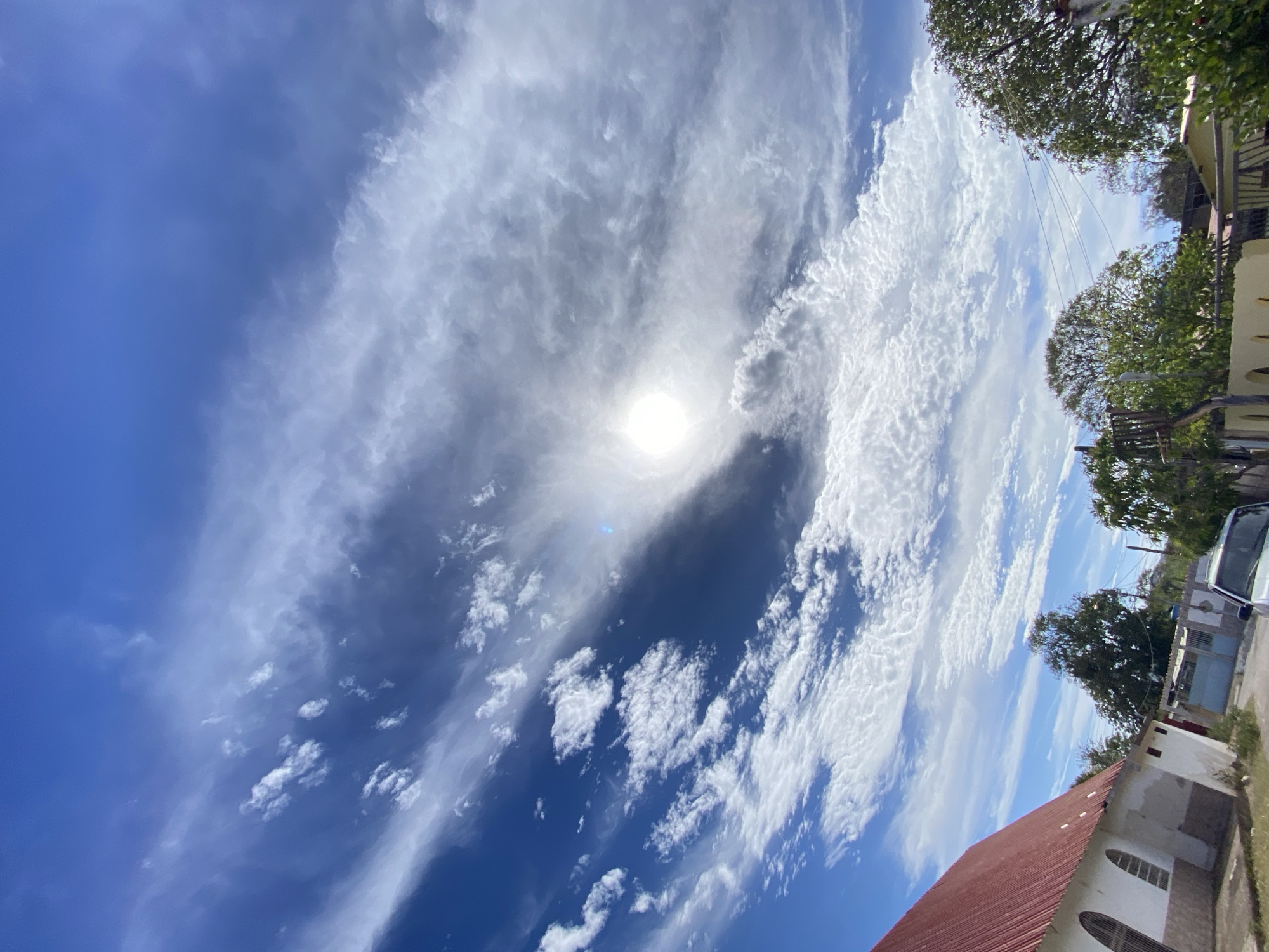The image, taken in Juan Griego, Venezuela, presents a view looking up at the sky with a bright sun visible through a layer of thin, scattered clouds. The sky transitions from deep blue in the lower-left corner to areas filled with elongated, streaky clouds extending across the frame. Along the lower-right edge of the image, a portion of a red-tiled roof of a building is visible, along with parts of the building's wall. Above the building, glimpses of trees and utility poles can be seen. A white car is also partially visible on the right. The environment appears to be a residential area on a sunny day. There are no people visible in the scene.