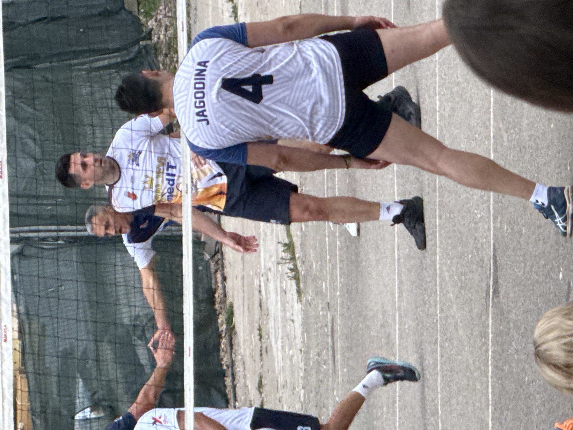 The image depicts a group of male volleyball players on an outdoor court, likely in Budva, Montenegro, given the context. In the foreground, a player wearing a white jersey with "JAGODINA" and the number "4" on the back is positioned with his legs spread, his gaze directed forward. To his left, another player in a white jersey with orange accents and "medIT" visible is in mid-action, looking to his right. Behind them, two players are shaking hands over the volleyball net, a common gesture of sportsmanship. The court surface appears to be asphalt with white lines marking boundaries. A green net stretches across the middle of the frame, separating the players. In the background, a dark fence or netting is visible, along with some greenery and a building structure. The overall setting suggests a daytime sporting event.