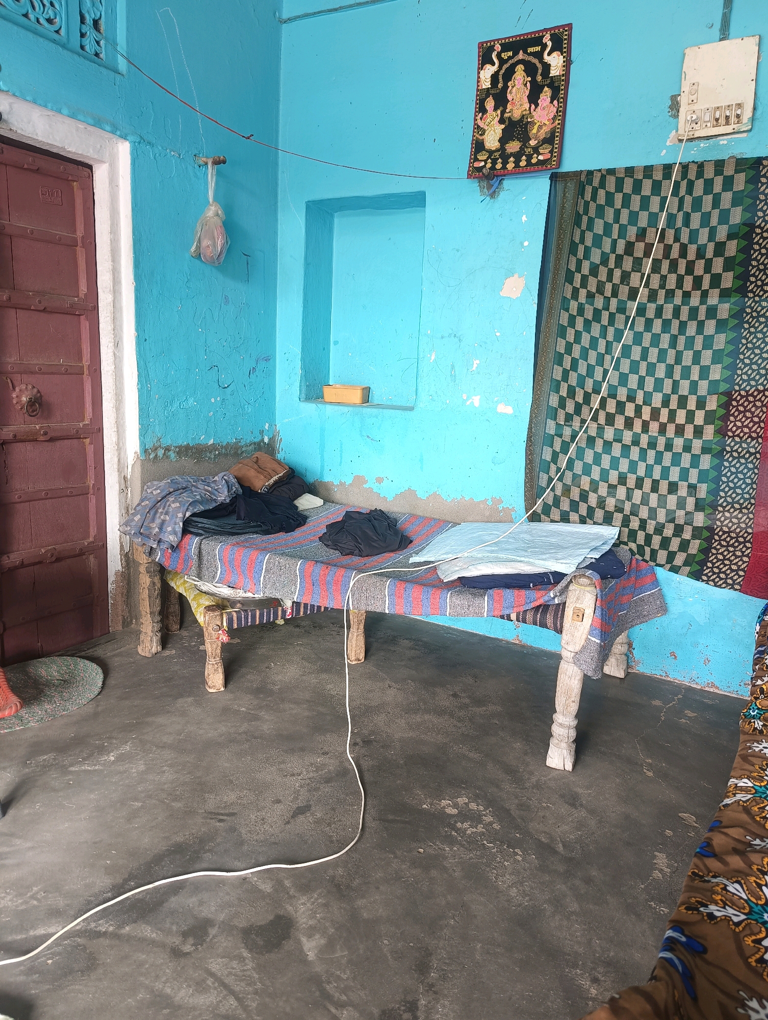 The image shows a rustic bedroom in Chhapri Bari, India. A wooden cot, draped with striped blankets and clothes, sits in the center of the room. A patterned curtain hangs on the right wall, next to a religious picture depicting Hindu deities. The walls are painted a vibrant blue, with a single door on the left side. A white electrical cord snakes across the floor. The room appears sparsely furnished, with a simple mat on the floor near the door. The overall impression is one of a humble dwelling.