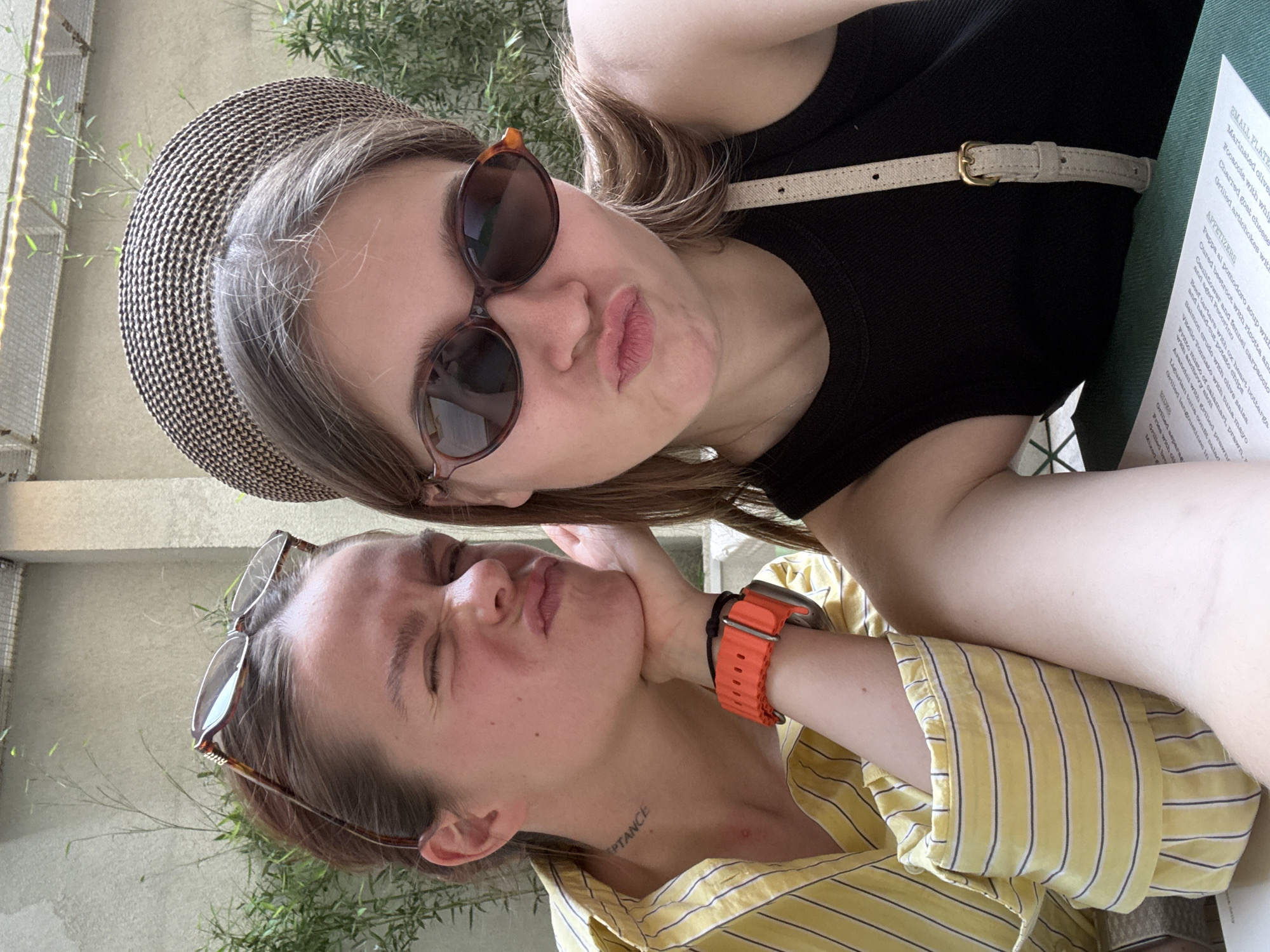 Two women are posing for a selfie at an outdoor restaurant. The woman on the top wears a black tank top, a tan hat, and brown sunglasses. The woman on the bottom wears a yellow and white striped shirt, orange watch, and has a tattoo on her neck. The background includes greenery, a metal fence, and a menu from the restaurant. Based on the text on the menu, it is likely that the restaurant is located in Belgrade, Serbia. The menu is titled in English, and the dishes listed have Italian names.