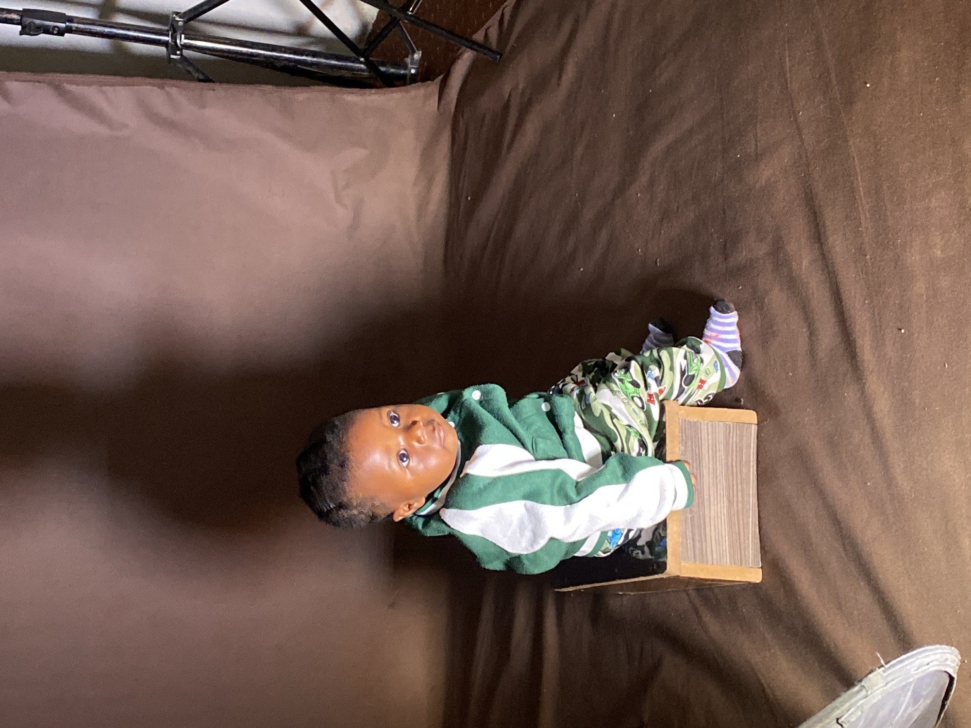 A young child, appearing to be a toddler or baby, is positioned in what looks like a makeshift indoor photo studio. The child has dark skin and hair, and is wearing a green and white striped long-sleeved top or jacket over patterned pants, possibly camouflage, and colorful striped socks. They are sitting or leaning against a small, light brown wooden box, positioned centrally in the frame. The child is looking directly towards the camera with wide, alert eyes and a neutral expression.
The background consists of a continuous brown fabric, draped to form both a vertical backdrop and a horizontal surface beneath the child. In the upper left corner, parts of a metallic stand or frame, likely for the backdrop or lighting equipment, are visible. Another object, possibly a light-colored shoe, is partially visible in the bottom right corner of the frame. The overall impression is a posed photograph. The image was taken in an indoor setting, and the provided context suggests the location is Vom, Nigeria.