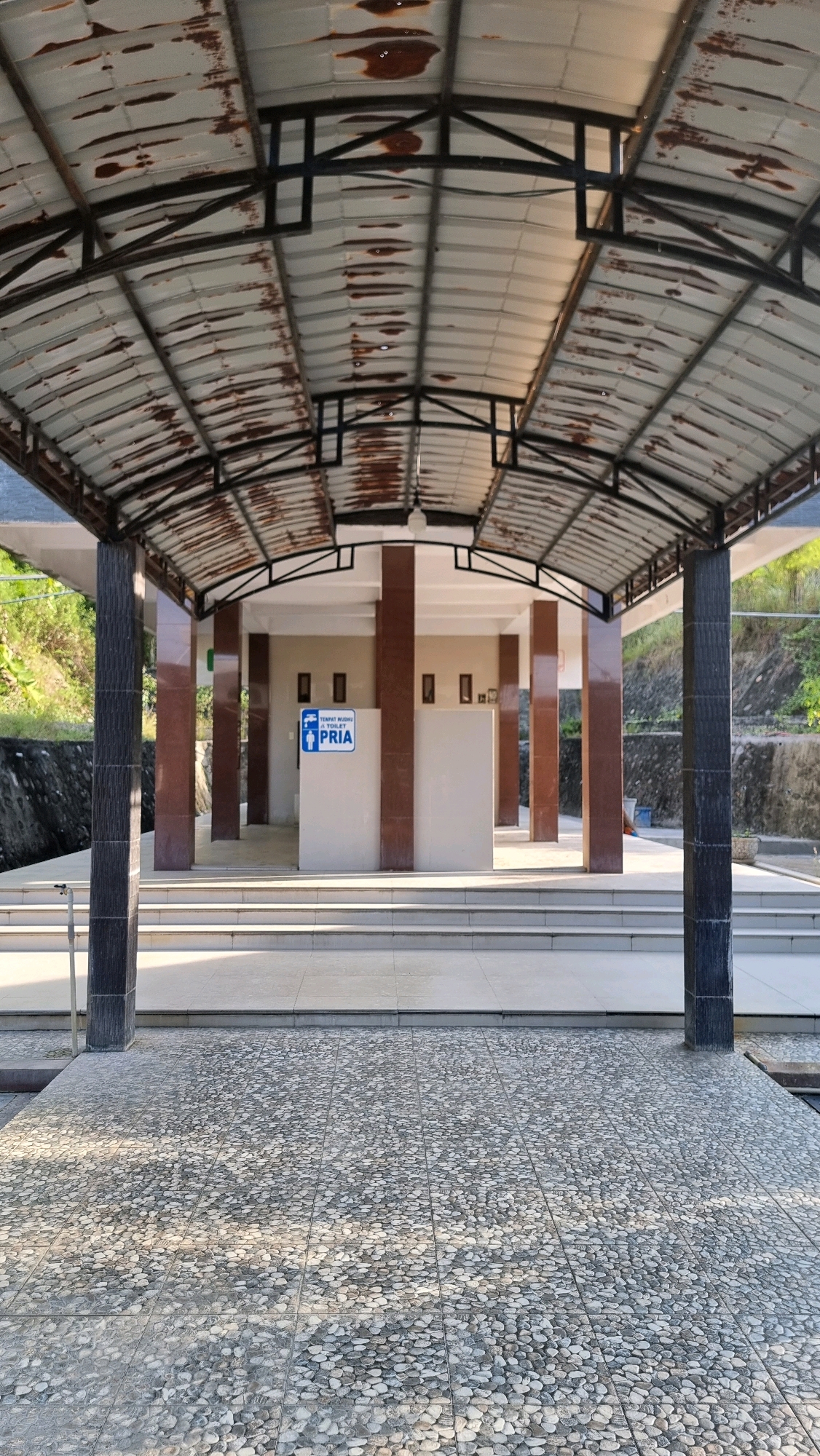 The image shows the entrance to a public restroom facility. The structure is covered by a weathered, rusted metal roof supported by dark grey and brown pillars. A sign with the word "PRIA" (Indonesian for men) and a male figure icon is visible on a white wall within the restroom area. The ground in front of the restroom is paved with small, greyish pebbles arranged in a tiled pattern, and there are several steps leading up to the entrance. The overall scene appears to be located in an outdoor setting, with vegetation and a stone wall visible in the background. The lighting suggests it is daytime, with sunlight casting shadows on the ground and steps. The facility seems to be situated in a public area, possibly a park or a rest stop, given the outdoor elements and the public nature of the restroom. The image does not depict any people or specific actions. The location is identified as Blangrambong, Indonesia.