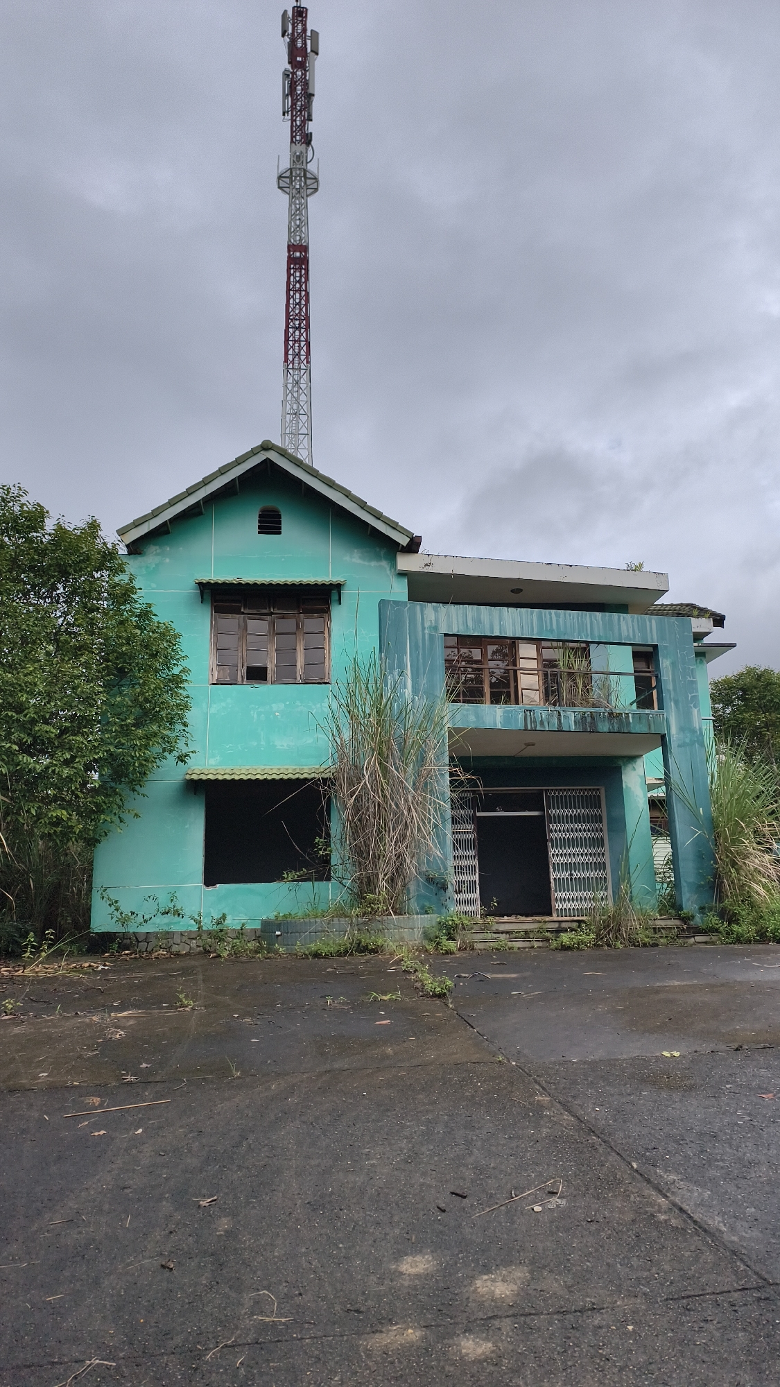 An abandoned, two-story building painted teal stands under a cloudy sky in Bờ Y, Vietnam, with a tall, red and white telecommunications tower visible behind it. The building's windows are broken or boarded up, and the facade is peeling. Overgrown vegetation, including tall grasses, surrounds the structure and encroaches on the cracked asphalt in front. The ground is damp, suggesting recent rain. A collapsible metal gate is visible at the entrance. The scene appears desolate and neglected.
