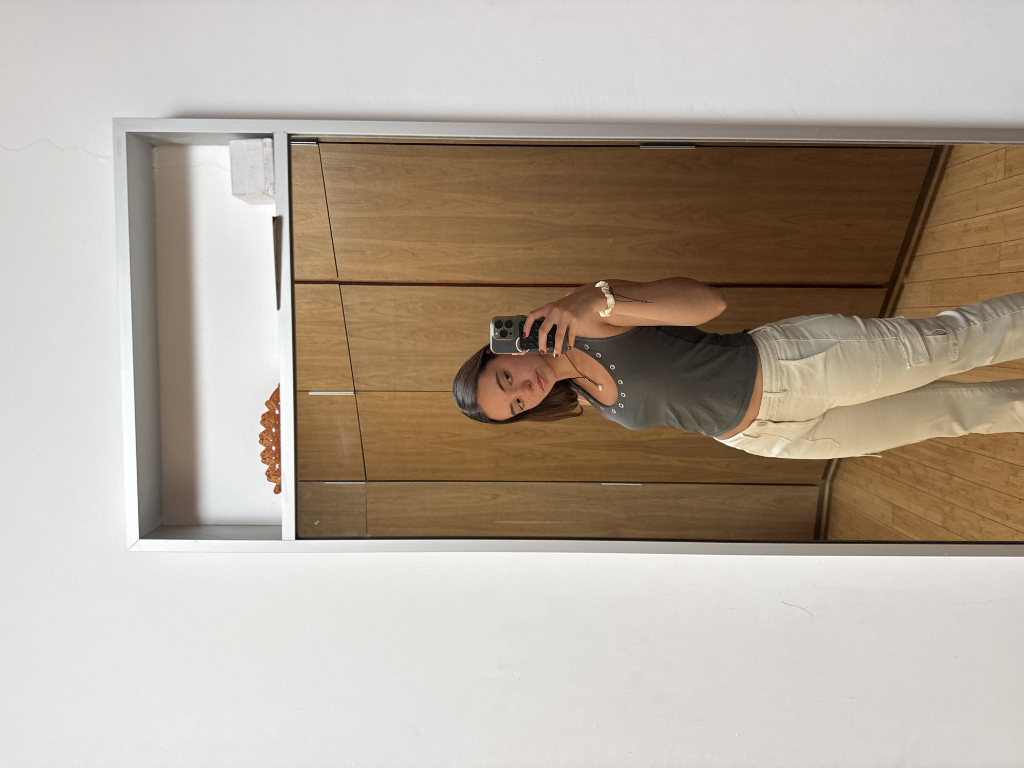 A young woman is taking a selfie in a mirror. The mirror has a silver frame and reflects her standing in front of a light wooden wall with horizontal panels. The reflection shows a hardwood floor, likely also made of light wood. The woman is wearing a dark gray tank top and light beige or cream-colored pants. She is holding a smartphone in her right hand, which is raised to capture the image. She also wears a light-colored bracelet on her right wrist. To the left of the mirror, there is a white wall with a decorative object placed inside a small, open shelf or cubby. The location appears to be indoors, potentially in a bedroom or dressing area. The city of San Bernardino Tlaxcalancingo, Mexico, cannot be inferred from this image.