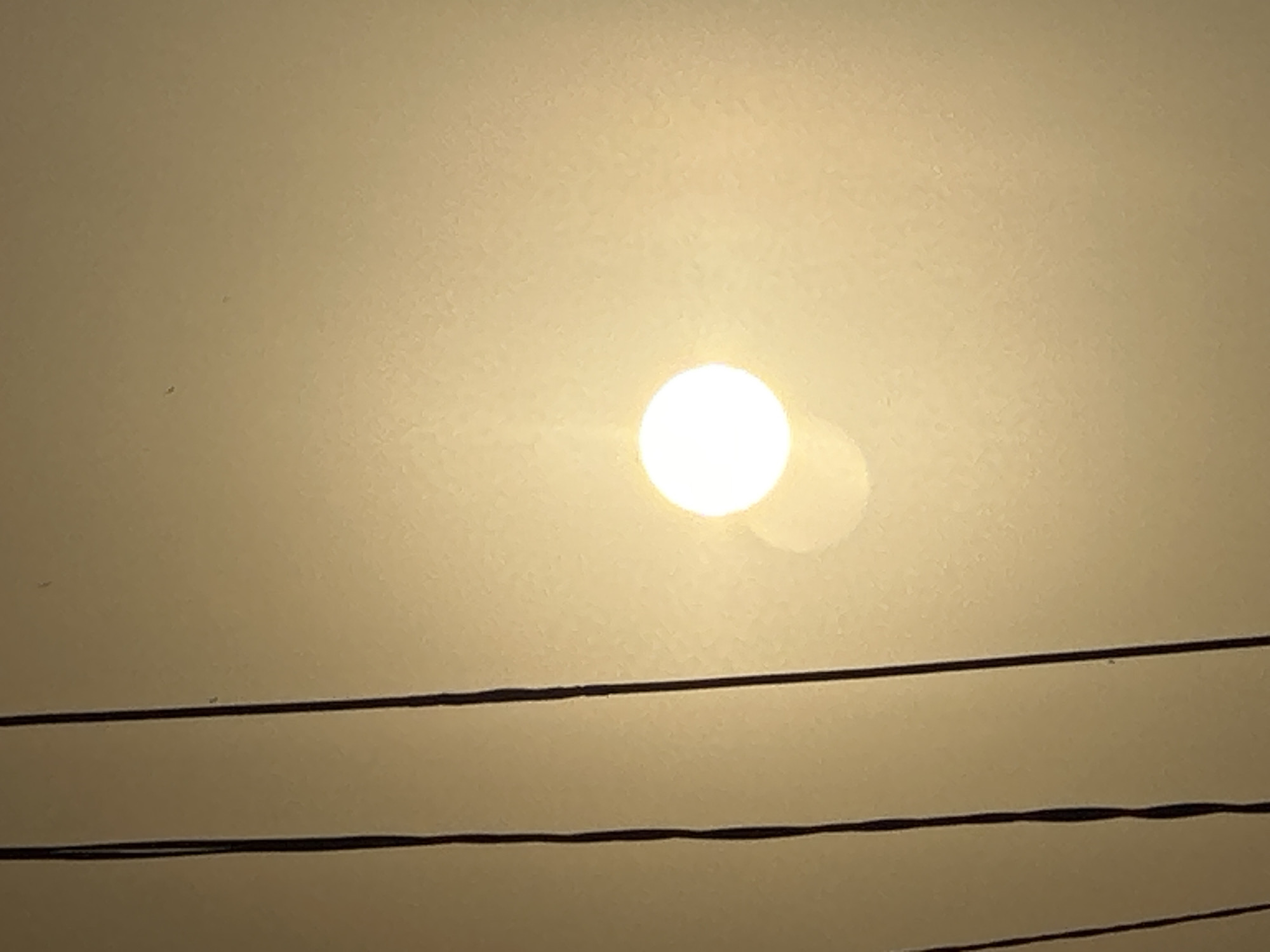 The image shows a bright, spherical object, likely the sun, obscured by a hazy or smoky atmosphere. The sky is a uniform pale yellow-gray. Three dark, horizontal lines, likely power lines or telephone wires, run across the lower portion of the frame. No people or other objects are visible. The location is unknown.