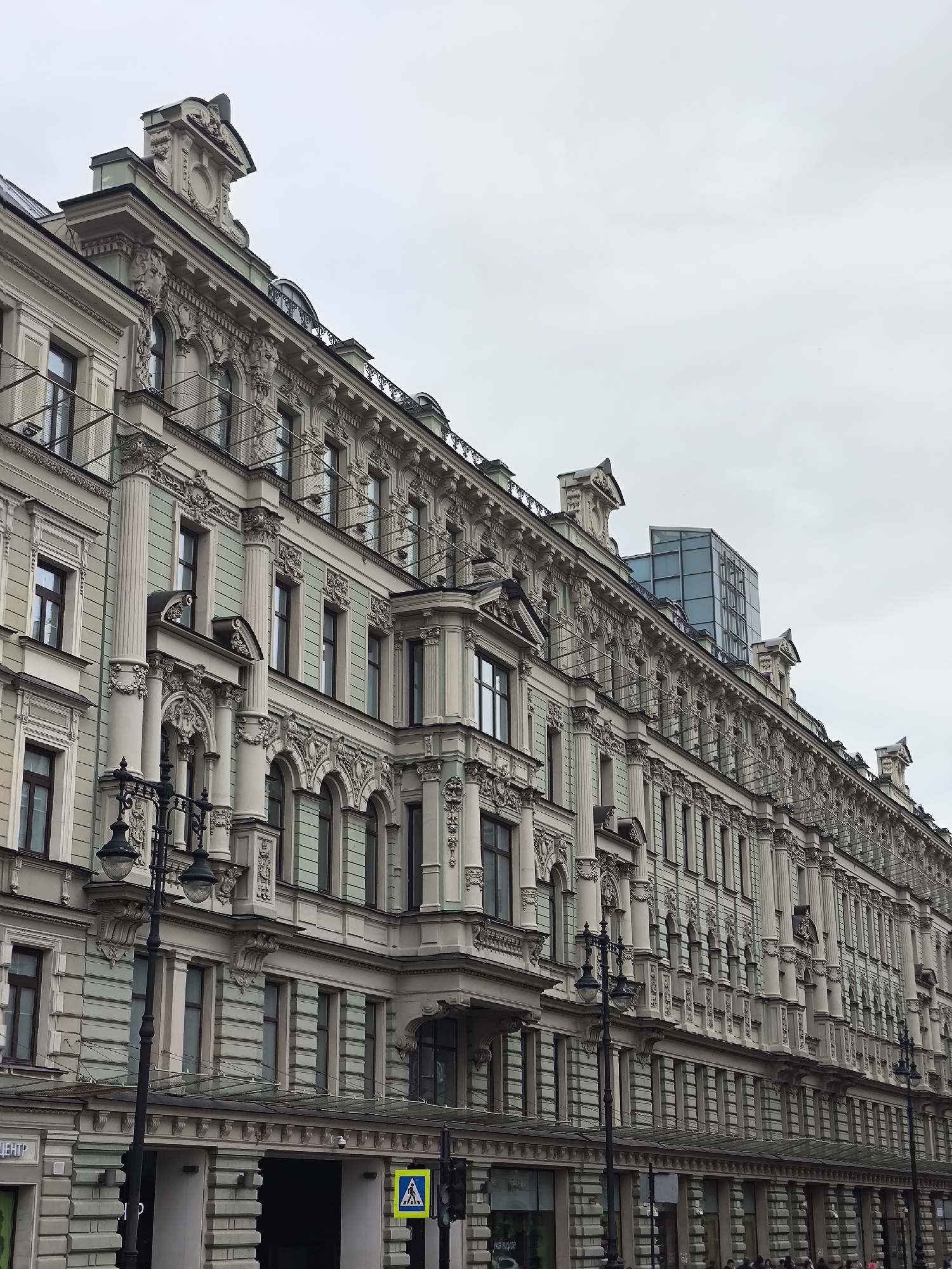 This is a low-angle, exterior, daylight photograph of a long, ornate, neoclassical building in Saint Petersburg, Russia. The building has a pale green and cream facade, adorned with intricate sculptural details, columns, and decorative moldings. The architectural style suggests it is a historic building. The windows are tall and rectangular, with some featuring small balconies or decorative elements. There are multiple streetlights in the foreground, typical of European cities. The sky is overcast, indicating a cloudy day. A modern glass structure is visible on the roof of the building, contrasting with the older architecture. In the lower portion of the image, there are storefronts and a pedestrian crossing sign. The scene appears to be a public street in a busy urban area.