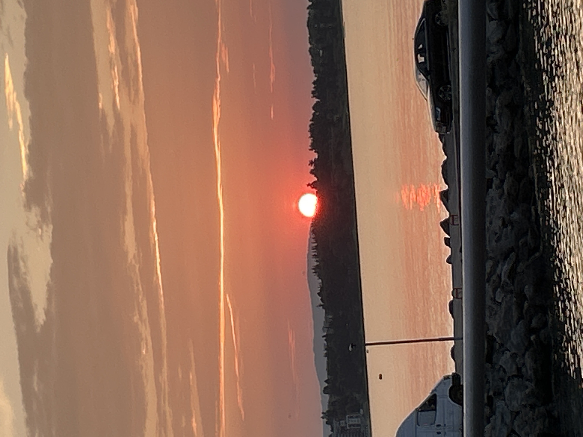 A horizontal, medium-shot photograph depicts a sunset over the sea in Ravda, Bulgaria. The sky is filled with bands of orange and pink clouds, with the setting sun a bright, reddish-orange orb positioned just above the tree line on the distant shore. The water in the foreground reflects the warm hues of the sunset, with a shimmering effect. Along the edge of the water, a rocky embankment is visible. On a paved path above the embankment, a white van is parked on the left side of the frame, and a dark-colored sedan is parked on the right. The tree line on the opposite shore is dark and silhouetted against the bright sky, with some buildings visible among the trees on the far left.