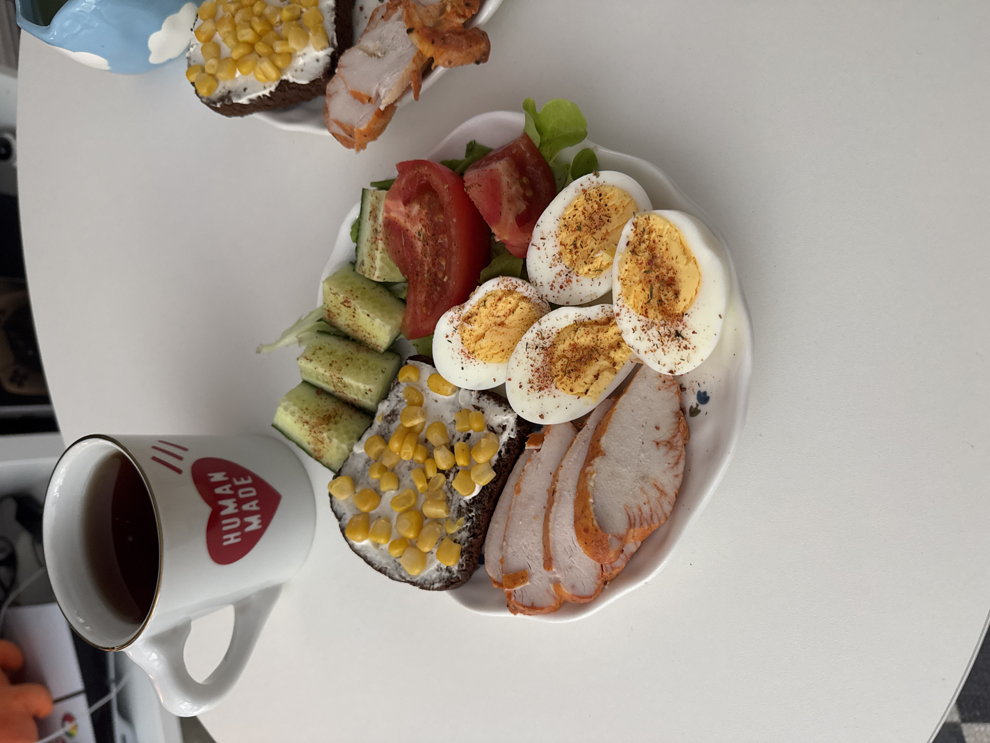 The image shows a breakfast laid out on a white table. The main subject is a plate of food, which includes sliced hard-boiled eggs sprinkled with spices, sliced cooked turkey or chicken, chopped tomatoes, cucumber slices, and a slice of dark bread topped with cream cheese and corn. To the left of the plate is a white mug with a red heart and the words "HUMAN MADE" printed on it, filled with a dark liquid, likely tea or coffee. In the background, another piece of bread with cream cheese and corn is visible, as well as some cooked meat. The scene appears to be an indoor setting, possibly a kitchen or dining area. The lighting suggests it could be daytime. The overall impression is of a home-cooked meal. The text visible is "HUMAN MADE" on the mug. The location is specified as Moscow, Russia.