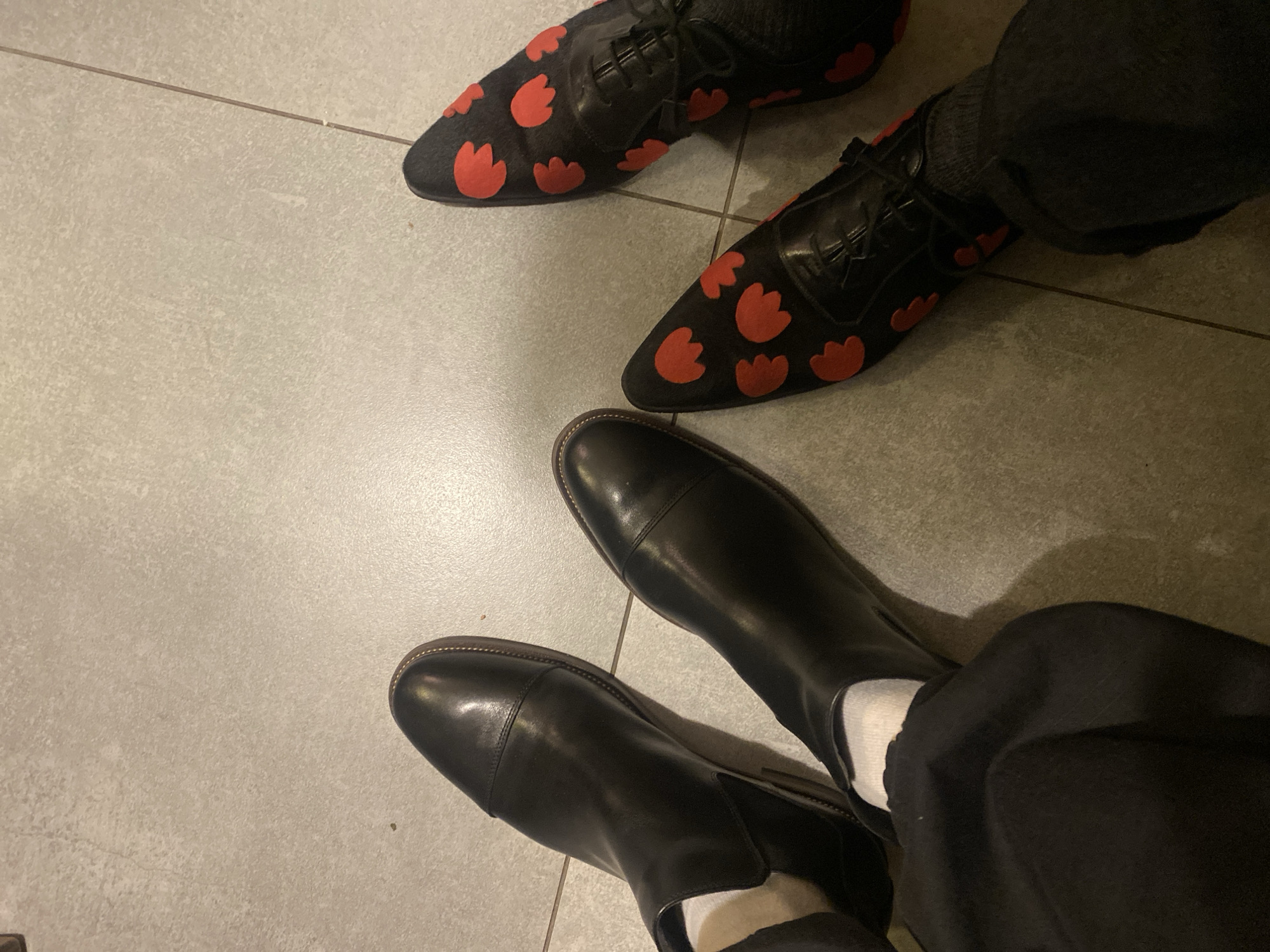 The image shows two pairs of shoes on a light gray tiled floor. The top pair consists of black leather lace-up shoes with red floral patterns, worn with dark gray socks. The bottom pair is comprised of black leather boots, worn with white socks and black trousers. The tile floor is consistently light gray and suggests an indoor setting. The location within Moscow, Russia, is not discernible from the visual information provided.