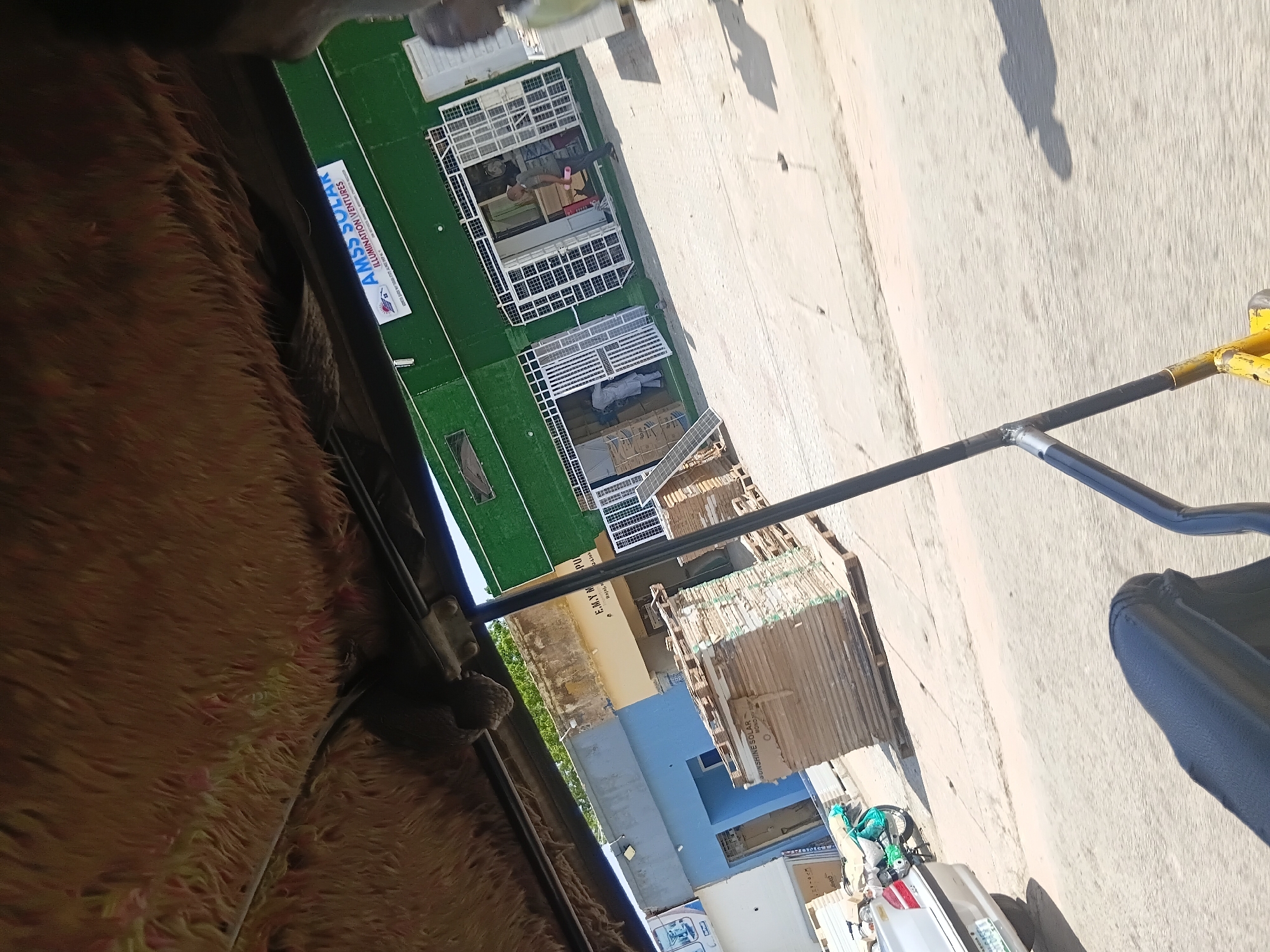 The image depicts a street view in Jalingo, Nigeria, likely taken from the interior of a vehicle. In the foreground, the fuzzy, brown interior of a vehicle's ceiling is visible, along with parts of the vehicle's structure. The middle ground features a row of shops and commercial buildings. The most prominent building is painted green and has white metal grates over its large glass doors and windows. A sign on this building reads "AMSS SOLAR ILLUMINATION VENTURES". To the left of this green building, there are other structures, including one painted blue. Piles of wooden pallets stacked with what appear to be solar panels or similar materials are placed outside the buildings. A white car is parked on the left side of the street, and a green motorcycle is parked beside it. People are visible inside and in front of the shops, with one person carrying a pink object. The ground is paved, and shadows suggest a sunny day. The overall impression is a commercial street scene in an urban environment.
