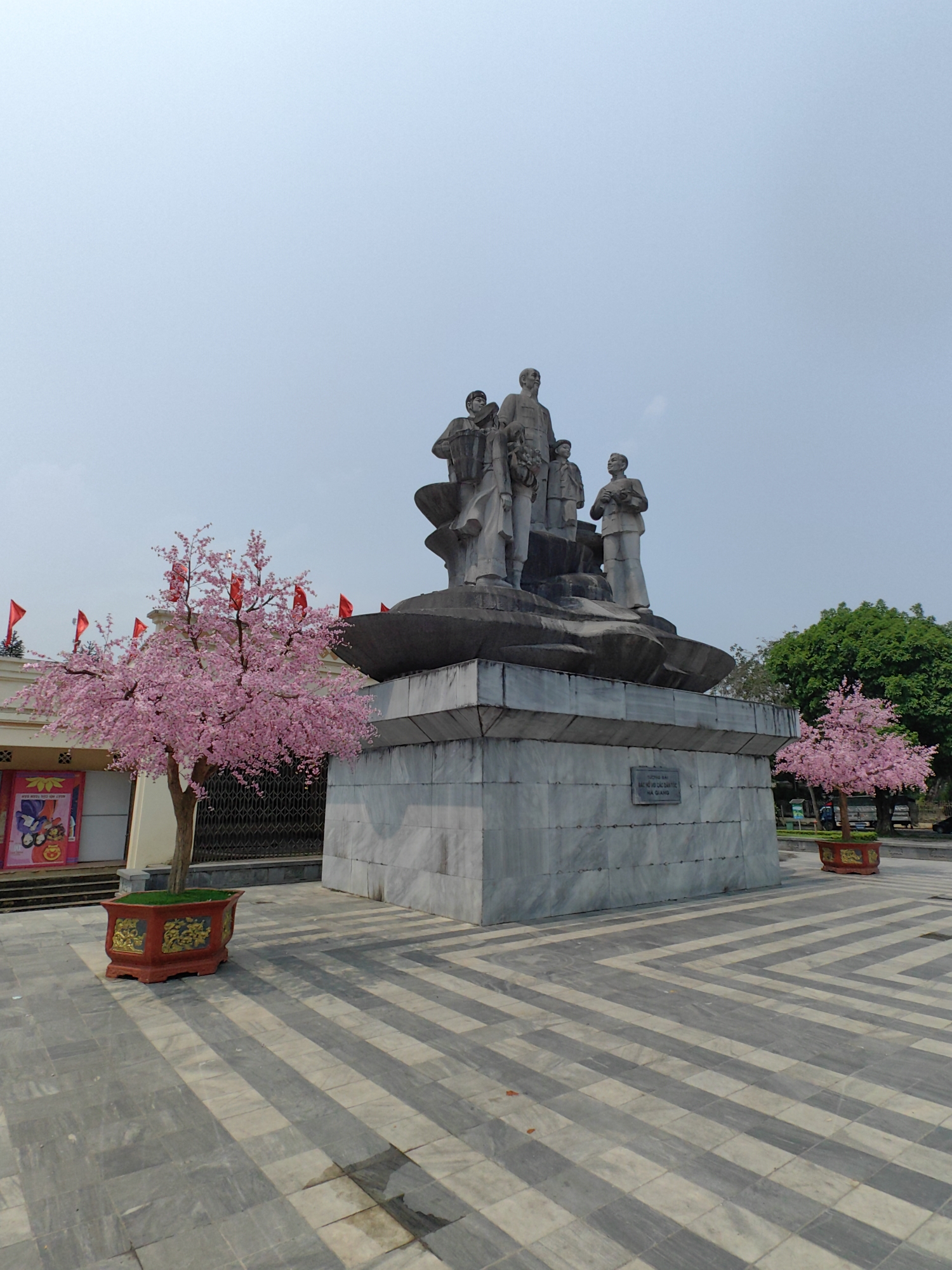 The image depicts a statue in Thành phố Hà Giang, Vietnam. The statue is a group of figures atop a gray, marble-like rectangular base. A sign is attached to the base. The statue is surrounded by paved ground with a striped pattern. Two artificial pink trees in decorative pots flank the statue, one on either side. In the background, buildings with red flags and green trees can be seen under a clear sky.