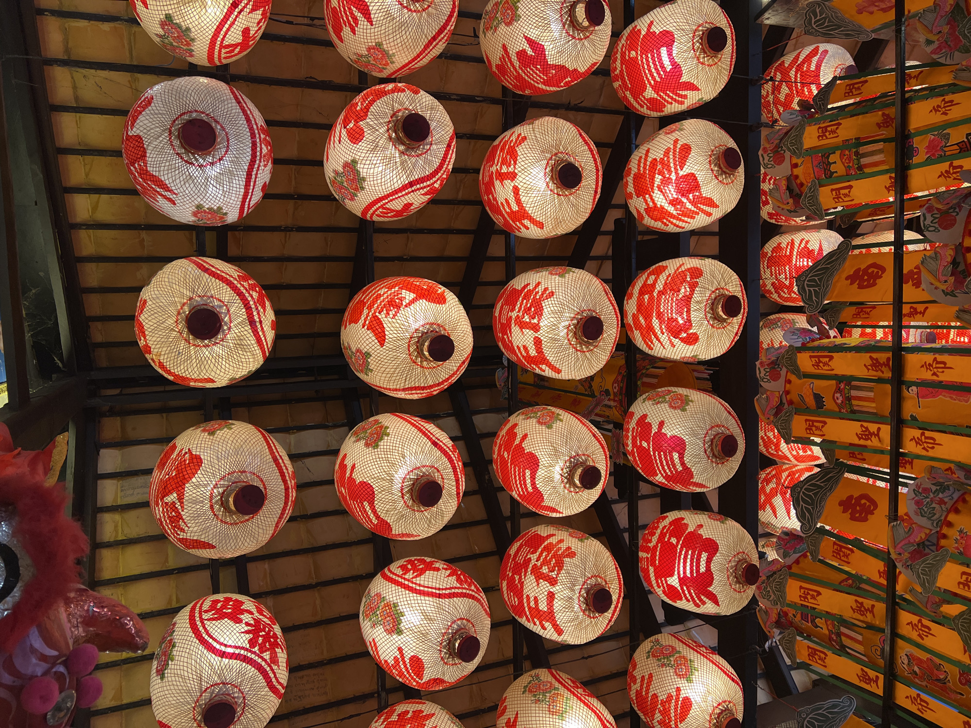 This image, captured in Baan Hua Thanon, Thailand, presents a high-angle view looking upwards at a dense arrangement of illuminated lanterns suspended from a ceiling. The primary subjects are numerous spherical or oblate spheroid lanterns, predominantly displayed in the left and central two-thirds of the frame. These lanterns are constructed from a translucent, light-colored woven material, possibly rattan or bamboo, and are internally lit, casting a warm yellowish-orange glow. Each lantern features distinct red stylized patterns, some resembling calligraphy or auspicious symbols, and several incorporate subtle pink and green floral motifs within the weave. A dark, cylindrical fixture is visible at the lower pole of each spherical lantern.
To the right, a different style of lantern is visible, appearing as multiple vertically hanging, rectangular or banner-like yellow panels. These yellow lanterns are adorned with prominent red Chinese characters and various pictorial elements, including figures in traditional attire and avian designs. They are arranged in tiered rows, suspended from a darker structural framework.
The ceiling background consists of dark-colored, possibly wooden, horizontal and vertical beams forming a grid pattern. Behind these beams, a lighter, yellowish-brown, textured surface is visible, likely the underside of a roof or an interior ceiling panel. In the bottom-left corner of the frame, a partial view of a red, furry decorative object with silver and pink accents is present, consistent with a portion of a traditional Chinese lion or dragon dance costume. No individuals are depicted.
