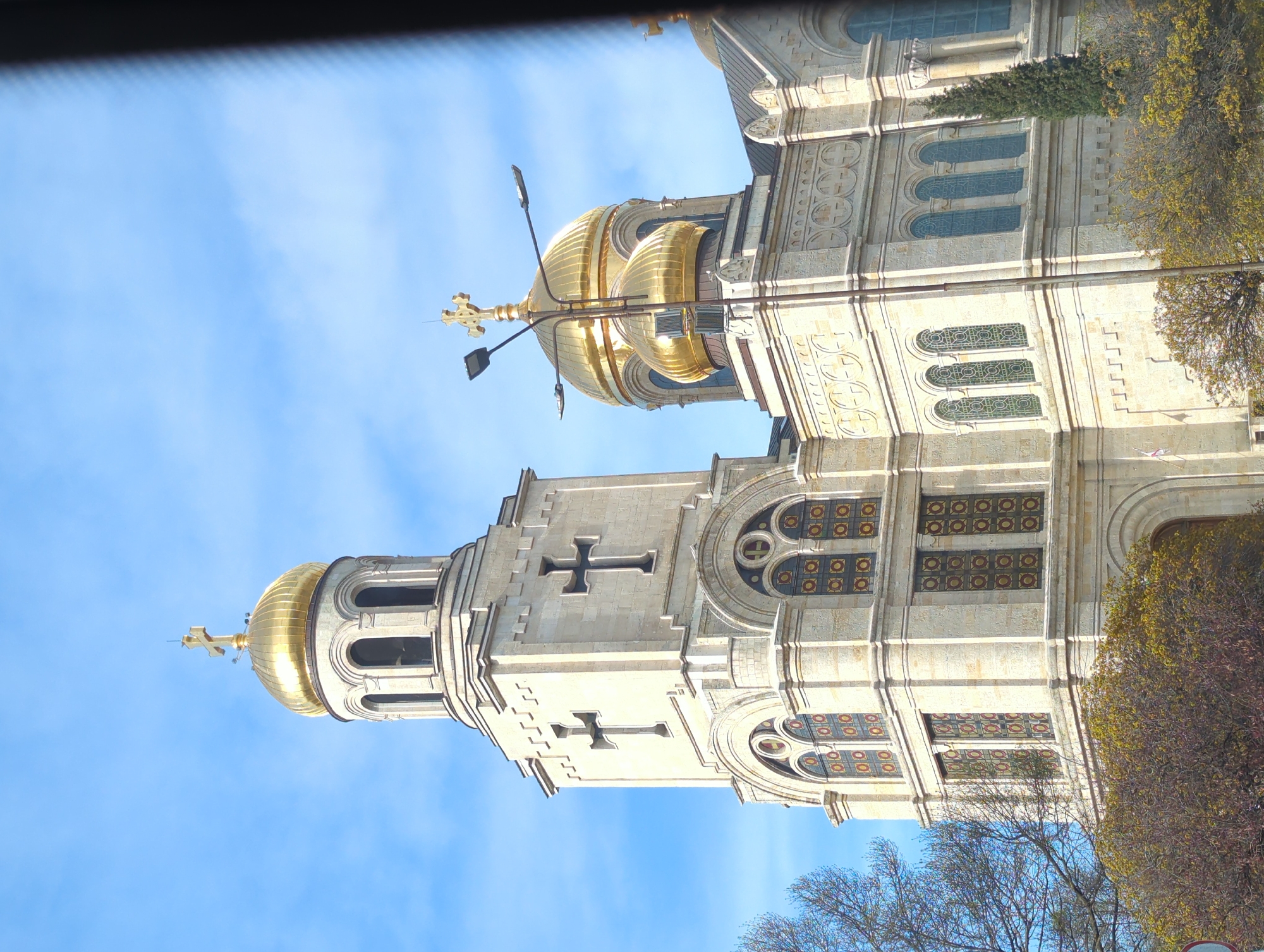 The image depicts the Dormition of the Mother of God Cathedral, a prominent landmark in Varna, Bulgaria. The cathedral is a large, ornate building with golden domes and crosses. The building is constructed from light-colored stone and features decorative carvings and windows. The domes are topped with gold crosses. The sky is blue with some light clouds. Trees are also visible in the foreground. There are street lights present.