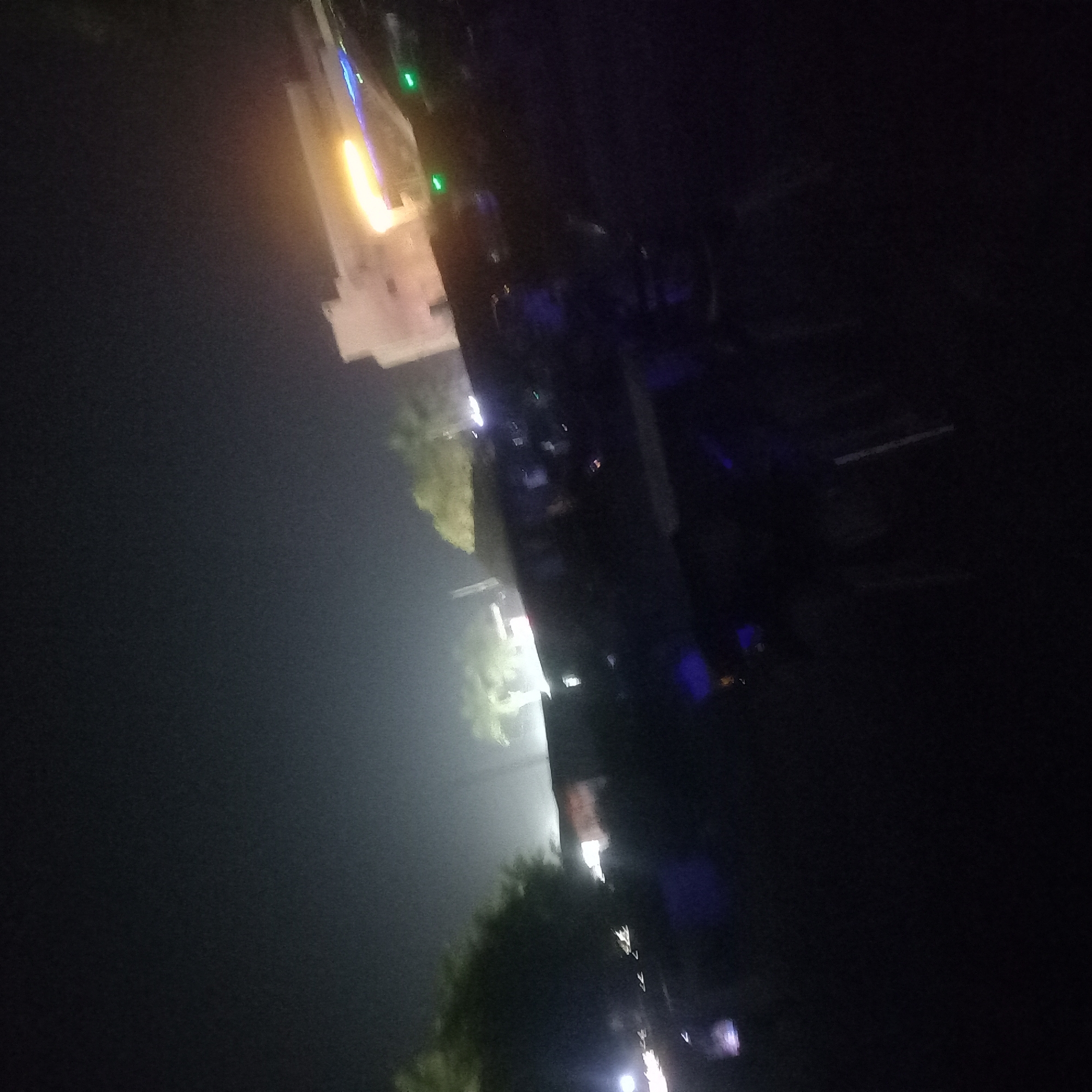 At night in Barden Yana, Nigeria, the image depicts a street scene under low light conditions. A building with lit windows, emitting a yellow glow, is visible in the upper left. A line of street lights extends along the street, with trees interspersed between the light sources. The foreground is dark, with indistinct objects. No people are discernible.