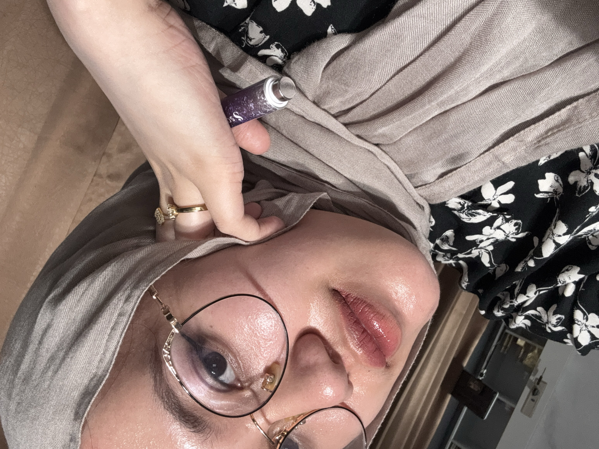 A person with light skin is holding a purple tube with a silver applicator to their face. The person is wearing gold-rimmed glasses and a light grey head covering. A gold ring is visible on their finger. The person is wearing a black and white floral patterned garment. The background includes a wooden floor and a glimpse of a curtain. This image was taken somewhere in Gadingrejo, Indonesia.