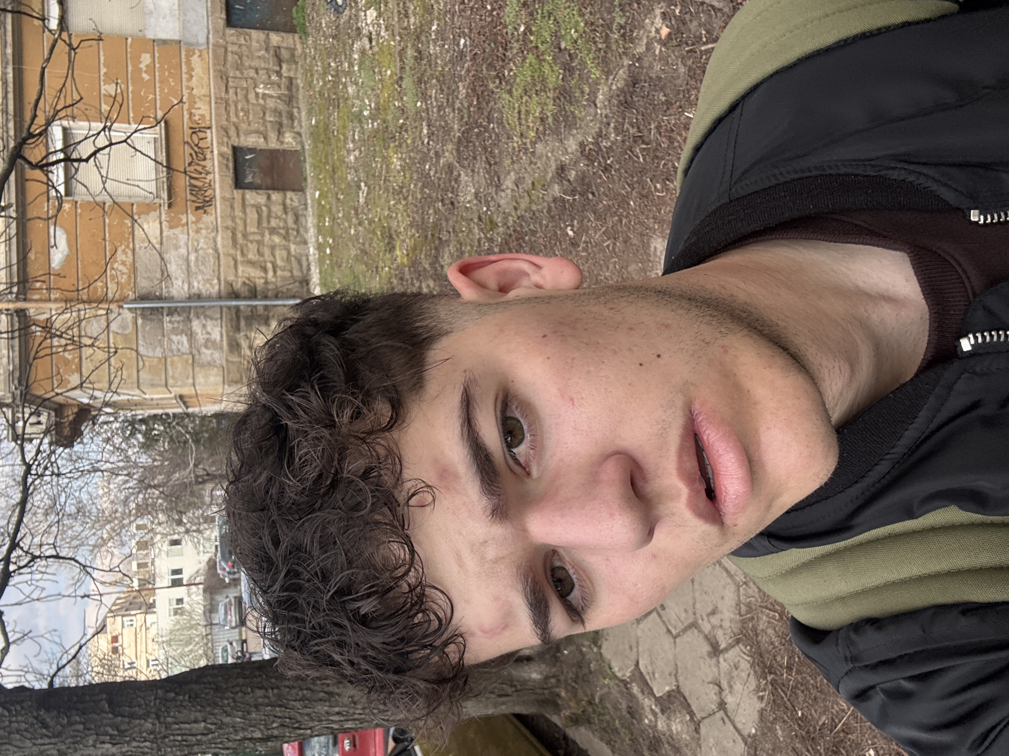 A young male subject occupies the foreground, positioned slightly off-center to the left, facing the camera directly. He has short, dark brown, curly hair with faded sides and prominent, dark brown eyebrows over dark brown eyes. His skin shows visible blemishes on the forehead and cheeks, and he has light stubble on his jawline and upper lip. His lips are slightly parted. He is wearing a dark brown long-sleeved shirt underneath a black bomber jacket with a ribbed collar. An olive-green padded backpack is visible over his right shoulder.
The background depicts an older, urban residential district in Sofia, Bulgaria. To the subject's left, a large deciduous tree with bare branches stands beside a section of irregular stone paving. Partially visible behind the tree is a red vehicle, likely a small hatchback, on a paved surface. To the subject's right, an aged, two-story building forms a significant portion of the background. Its lower half features a stone block foundation, while the upper floor is covered in peeling, orange-yellow stucco. A single window with closed horizontal blinds is visible on the upper floor, with dark graffiti, partially illegible, applied to the wall beneath it. At ground level on the building's right side, a dark, recessed opening, possibly a boarded-up door or window, is present. The ground in front of this building is a mix of sparse, dry grass and bare earth. In the distant background, beyond the trees and other foreground elements, several multi-story residential buildings are visible under an overcast sky.
