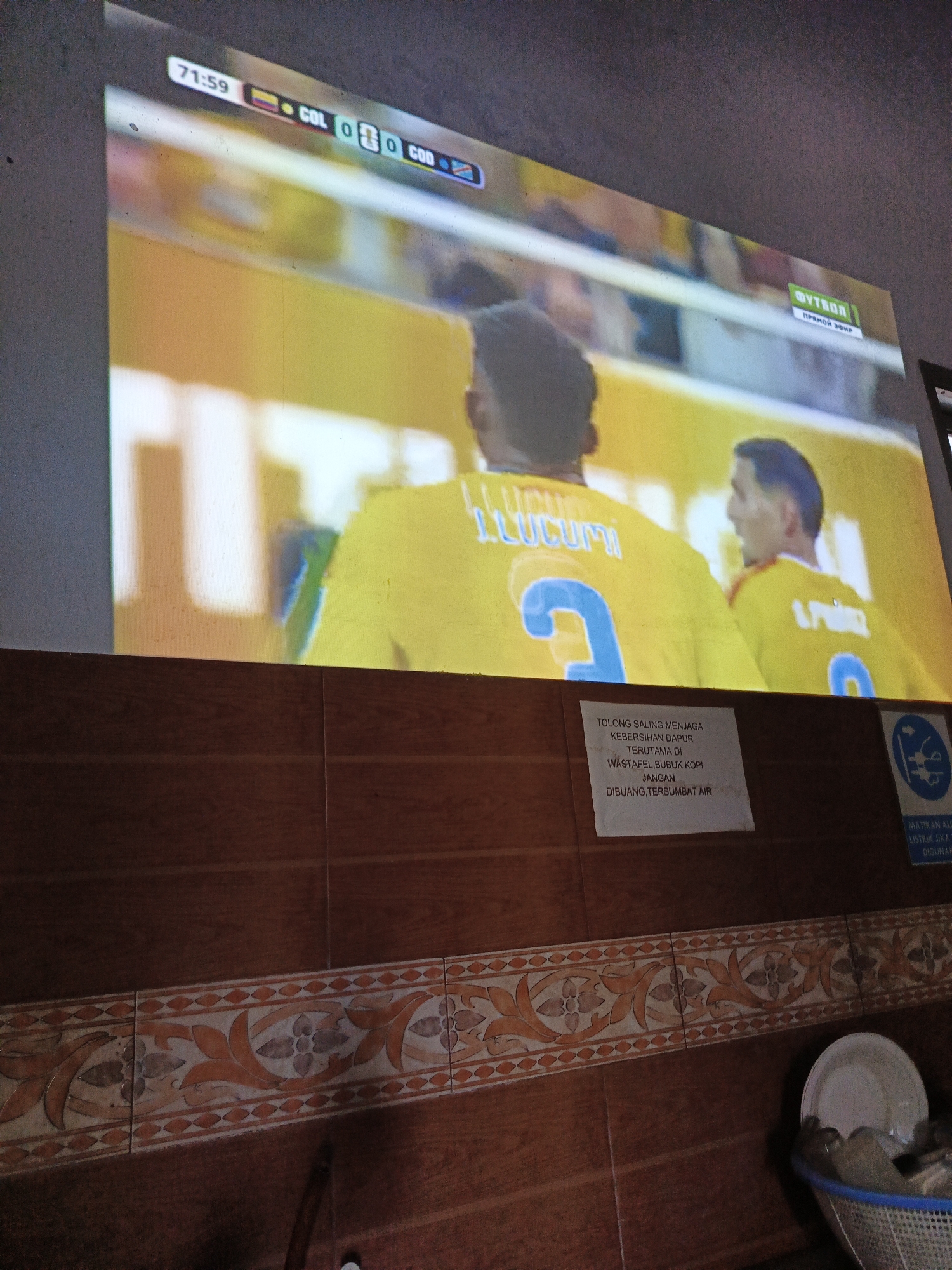 The image shows a projection of a soccer match on a large screen, likely in a public space or a home entertainment setup in Julicotmeurak, Indonesia. The score displayed on the screen is 71:59, with "COL" and "COD" represented by their respective country flags, indicating a match between Colombia and possibly another nation whose flag is not clearly visible but starts with "COD". The players on the screen are wearing yellow jerseys. Below the screen, there's a wall covered with wood-patterned tiles and a decorative border. A white sign with Indonesian text is affixed to the wall, reading "TOLONG SALING MENJAGA KEBERSIHAN DAPUR TERUTAMA DI WASTAFEL. BUBUK KOPI JANGAN DIBUANG,TERSUMBAT AIR." (Please maintain kitchen cleanliness, especially at the sink. Do not throw coffee grounds, it will clog the drain.) To the right of this sign, another blue sign with a pictograph indicating electrical safety is visible, and further to the right, a white plate is placed in a blue plastic basket filled with what appears to be kitchen items. The overall scene suggests a casual setting where people might gather to watch sports, with an emphasis on maintaining cleanliness in a shared kitchen space.