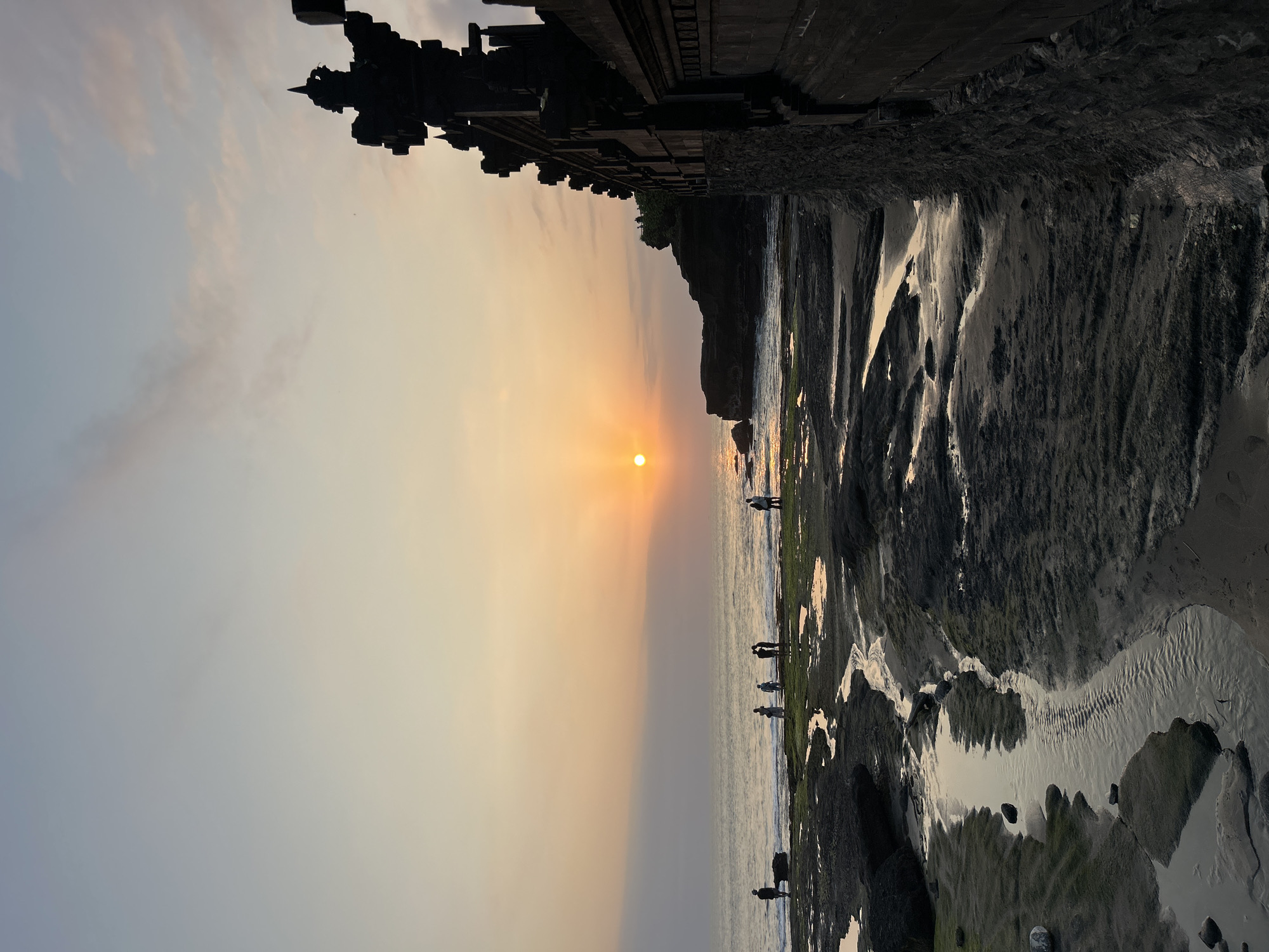 This image depicts a sunset scene at Tanah Lot Temple, a famous Hindu sea temple on the coast of Bali, Indonesia, not Kediri. Silhouetted figures are visible on the rocky shoreline, some standing near the water and others closer to the temple structure. The sun is low on the horizon, casting a warm glow over the ocean and the sky. The foreground shows a rocky beach with tide pools and a narrow channel of water. To the right, the distinctive architecture of Tanah Lot Temple is visible, with its dark stone structure and ornate detailing. The overall atmosphere is serene and picturesque.
