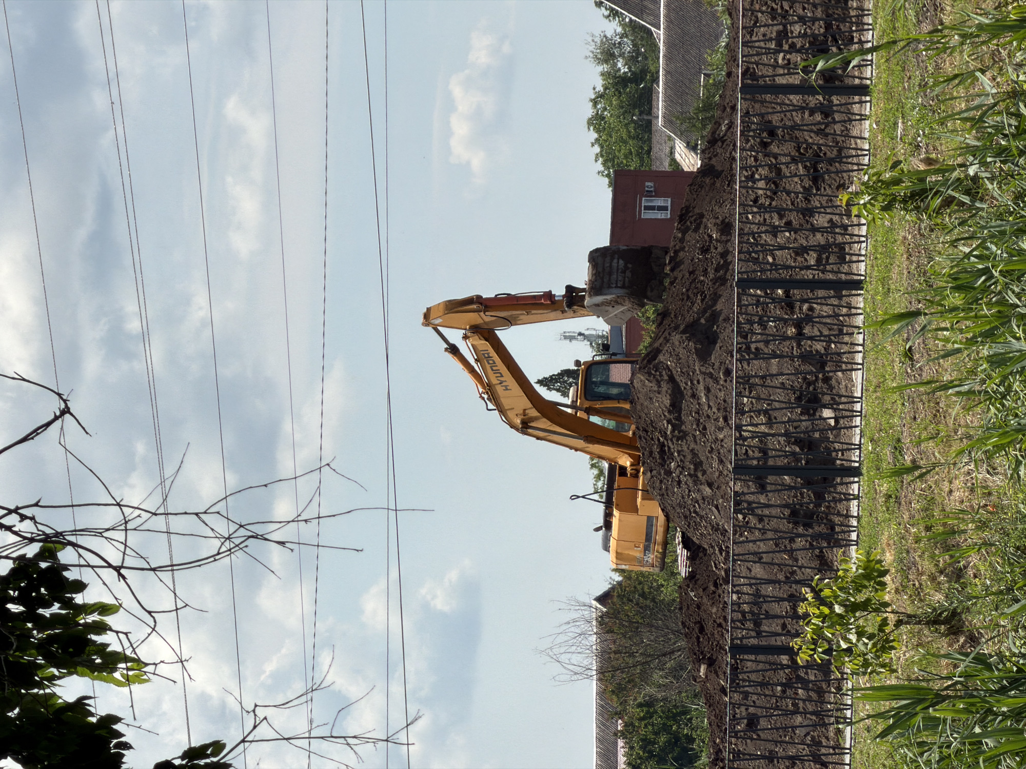 The image depicts a construction site in Leningradskaya, Russia. In the foreground, green vegetation and tall grasses grow along a paved path. Behind the path, a dark metal fence with a unique angled bar pattern separates the foreground from a large pile of dark soil.
Dominating the middle ground is a yellow Hyundai excavator, actively engaged in moving the soil. The excavator is positioned on top of the soil pile, with its boom extended and bucket lowered, appearing to be in the process of scooping or dumping earth. The tracks of the excavator are visible on the soil.
In the background, several buildings with pitched roofs are visible, some appearing to be residential. One building is a reddish-brown color with a white-framed window. The sky is partly cloudy, with scattered white clouds against a pale blue backdrop. Overhead, thin power lines are strung across the frame, with some branches of a leafy tree visible in the upper left corner. The overall scene suggests ongoing construction work in a semi-urban or suburban environment.