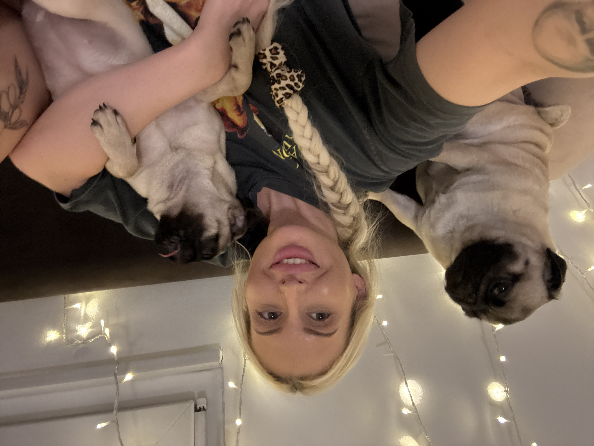 The image is an upside-down, close-up, selfie-style photograph featuring a woman and two pugs. The woman has blonde hair styled in a braid and is wearing a dark t-shirt. One pug is lying on her chest, with its paw resting on her arm, while the other pug is positioned in front of her face, with its head tilted to the side and tongue slightly out. The background is dimly lit, with a string of warm white fairy lights visible, suggesting the photograph was taken indoors at night or in a dimly lit room. The woman appears to be smiling and looking at the camera, while the pugs' expressions are difficult to discern due to the lighting and their positions. There are no visible objects other than the subjects and the fairy lights, and no clear indication of the location beyond an indoor setting. The overall mood of the photograph seems to be casual and affectionate, with the woman embracing her dogs.