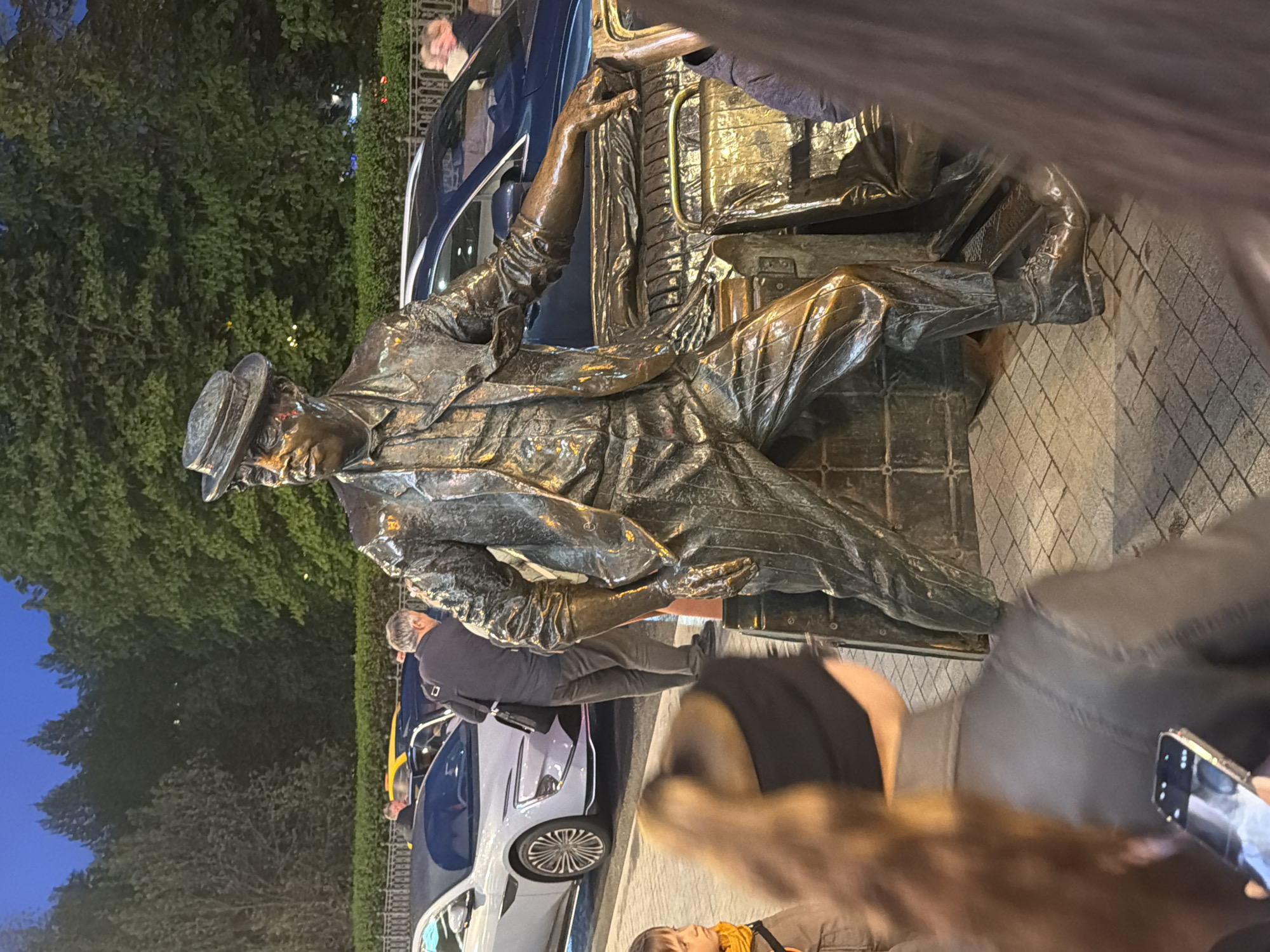 The image depicts a bronze statue of a man seated on luggage, wearing a hat and suit, seemingly a traveler. The statue is placed on a paved surface with a crosshatch pattern. Behind the statue are parked cars and lush green trees, indicating an urban environment. People surround the statue, some appearing to interact with it. A hand holding a smartphone is visible in the foreground, suggesting someone is taking a photograph. The setting is Moscow, Russia.