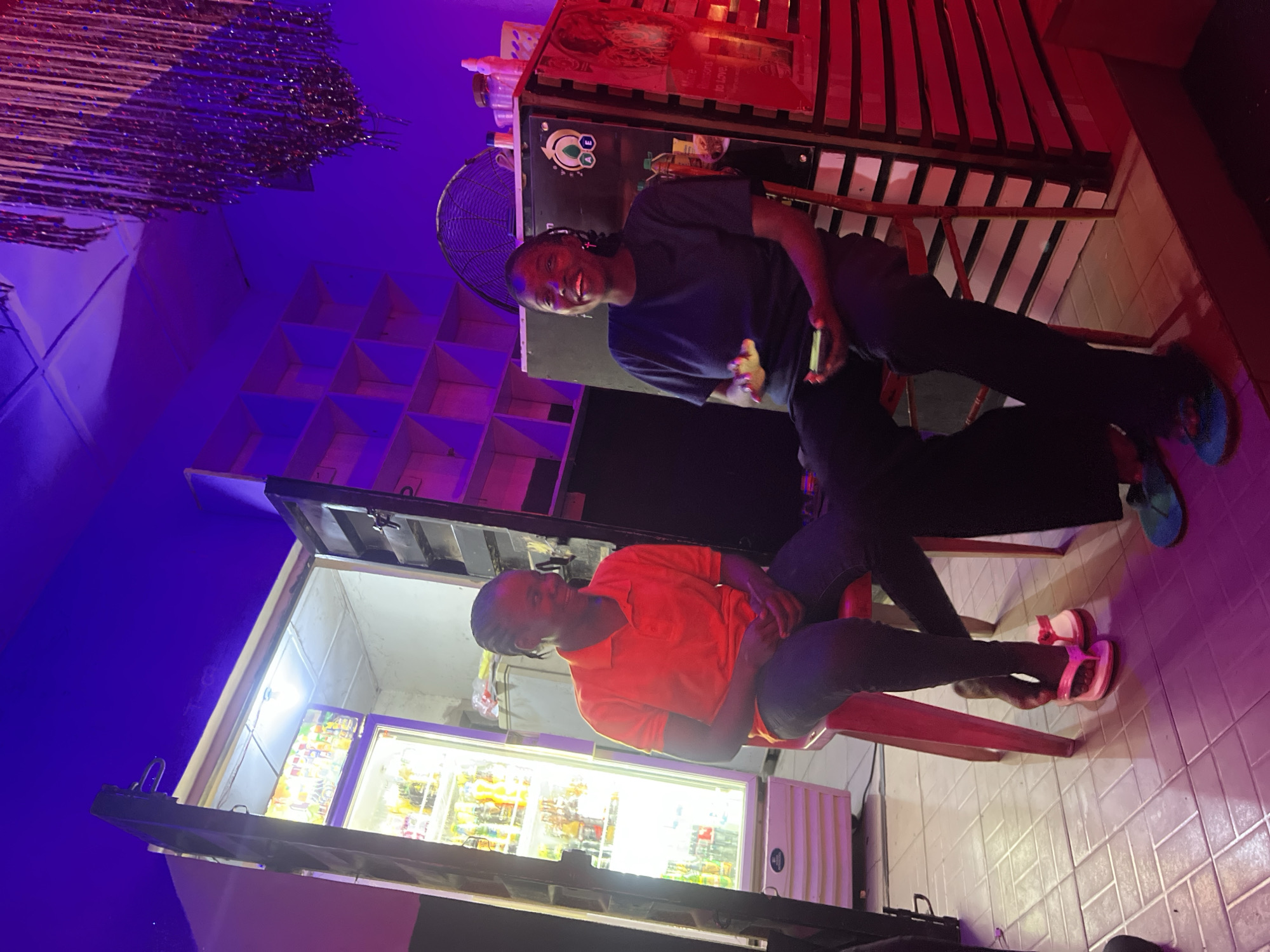 The image displays an indoor scene at a venue in Howayi, Nigeria. Two individuals, both appearing to be women, are seated on red stools, engaged in an interaction. The environment is illuminated by artificial lighting, casting prominent purple and red tones across the space, contrasted by bright white light emanating from a beverage cooler and an overhead bulb.
The individual on the left is wearing an orange polo shirt and dark trousers. Her dark hair is styled in braids, and she is observed smiling while looking towards the person on the right. Her hands are clasped in front. She wears pink flip-flops. Her right foot is positioned near the left foot of the second individual.
The individual on the right is wearing a dark t-shirt and dark trousers, with dark braided hair and small cross-shaped earrings visible. She is smiling broadly, holding a smartphone in her left hand, and her right hand is slightly raised. She wears teal-colored flip-flops. Her left foot is extended, making contact with the right foot of the individual on the left.
The background on the left features a tall, white, upright beverage cooler with a glass door, stocked with various bottled and canned drinks, emitting a bright white light. Above this cooler, an open section of a wall or serving hatch reveals an exposed lightbulb. The floor throughout the visible area consists of light-colored tiles. On the right side of the frame, a dark structure, potentially a counter, is present, topped with various containers and bottles. A round electric fan is positioned behind the right individual, resting on this structure. Further right, a section of a red-paneled wall is visible. Above the fan, a shelving unit with multiple open compartments is mounted on the wall. A decorative element, resembling metallic tinsel or strands, hangs from the ceiling in the upper right quadrant of the image.