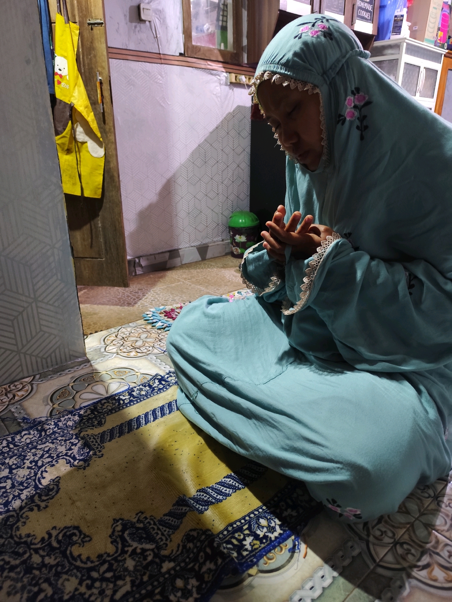 In Jambewangi, Indonesia, a person wearing a light blue prayer garment with floral embroidery is kneeling on a prayer mat. The prayer mat has a yellow base with intricate blue and white patterns. The surrounding environment includes a tiled floor, a wooden door, and a wall with a geometric pattern. A yellow apron with a cartoon bear is hanging on the door. The person's hands are clasped together in front of their chest. The lighting suggests an indoor setting, possibly a home.