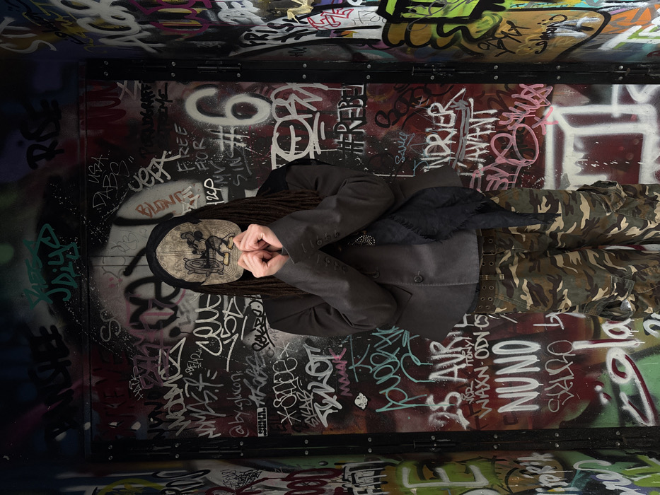 An individual stands centered against a heavily graffitied vertical surface in an urban setting, identified as Paris, France. The person wears a beige baseball cap featuring a distressed graphic of Mickey Mouse, from which dark dreadlocks emerge and frame the face. The individual's face is obscured by their hands, clasped together at chest height with interlocked fingers covering the nose and mouth area. They are dressed in a dark, likely charcoal grey, single-breasted blazer over a darker inner garment, accessorized with a dark, frayed-edge scarf draped around the neck and a metallic chain necklace. A dark belt with multiple eyelets is visible at the waist, securing camouflage-patterned cargo pants with prominent side pockets. The background is a dense collage of overlapping graffiti tags, words, and abstract markings in various colors including black, white, red, green, pink, and blue, applied to a dark metal or wood surface that appears to be a door or panel structure. Visible graffiti elements include "25 AVR," "NUNO," "By.Glacon," "FREE FOR MY SIN," and the number "6."