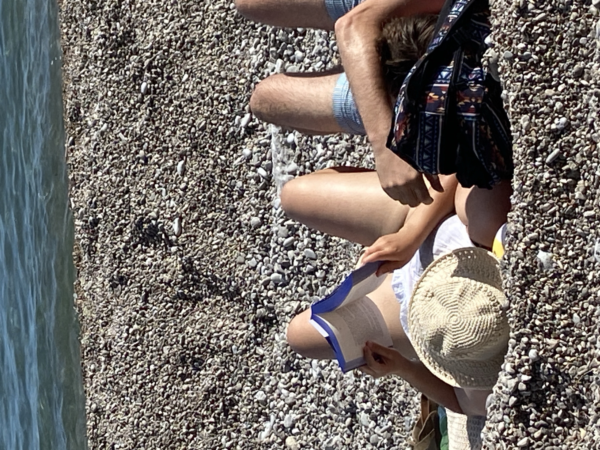 The image depicts a sunlit beach scene. The left side shows the edge of a body of water, possibly the sea, and the rest of the image is composed of a rocky beach. The image captures two people sitting on the beach. One individual is wearing shorts. The other person, who is wearing a white shirt and straw hat, is reading a book. A patterned bag is located near the first person. The beach consists of small, smooth rocks. The location is not explicitly identifiable as being in Prijevor, Montenegro from the image itself.
