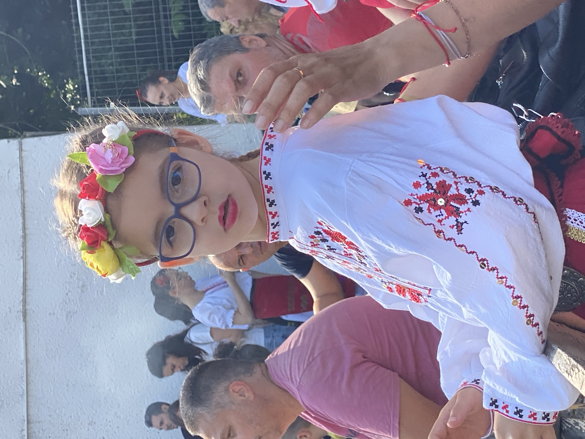 The scene is set outdoors in the city of Pleven, Bulgaria, under bright daylight. The primary subject is a young girl, estimated to be 8-12 years old, positioned in the foreground and slightly to the left of the central axis. She has light brown, wavy hair adorned with a floral headband featuring artificial blossoms in red, yellow, pink, and white. She wears blue-framed glasses and red lipstick. Her attire consists of a white traditional embroidered blouse with intricate red and black geometric patterns, including gold sequin accents, visible on the collar, chest, and sleeves. She faces the camera directly with a neutral expression.
Behind the girl, a diverse group of individuals is present. To her immediate left, a man with short grey hair, wearing a pink t-shirt, is visible from the side. Further left, partially obscured, are a man with dark hair and sunglasses, and a woman in a white t-shirt looking downwards. Another woman, further back, wears a white blouse and a red skirt.
To the right of the primary subject, a man with grey hair, wearing a dark t-shirt, looks towards the camera. His raised right hand, displaying a simple gold band on the ring finger, partially obscures his face. An arm, distinct from this man's, extends from the right edge of the frame, adorned with red string and beaded bracelets, resting above a visible black bag. Further in the background, a woman with dark hair pulled back, wearing a white shirt, is visible smiling behind a metal fence. Another man with grey hair is partially visible to the far right.
The background on the left consists of a plain, light-colored wall. Green foliage and trees are visible behind the fence on the right. A wooden beam or railing is present in the lower central foreground.