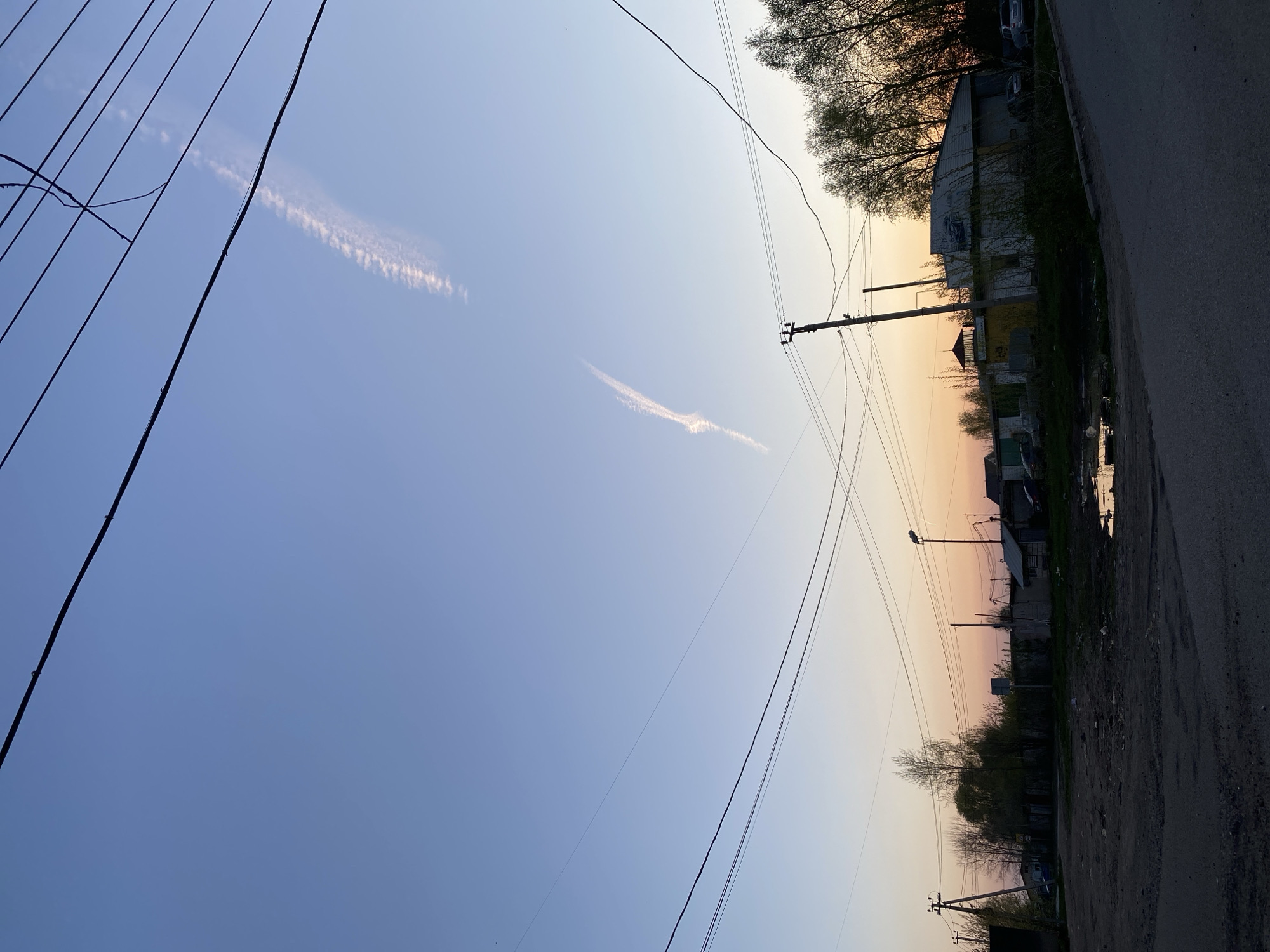 The photograph depicts a scene in Chaikovsky, Russia, at what appears to be dusk or dawn, based on the sky's color gradient from blue to orange. The perspective is from ground level, looking up at an angle. Power lines crisscross the sky. Two contrails are visible in the blue portion of the sky. The right side of the frame shows a street with buildings, trees, a lamppost, and puddles on the road.