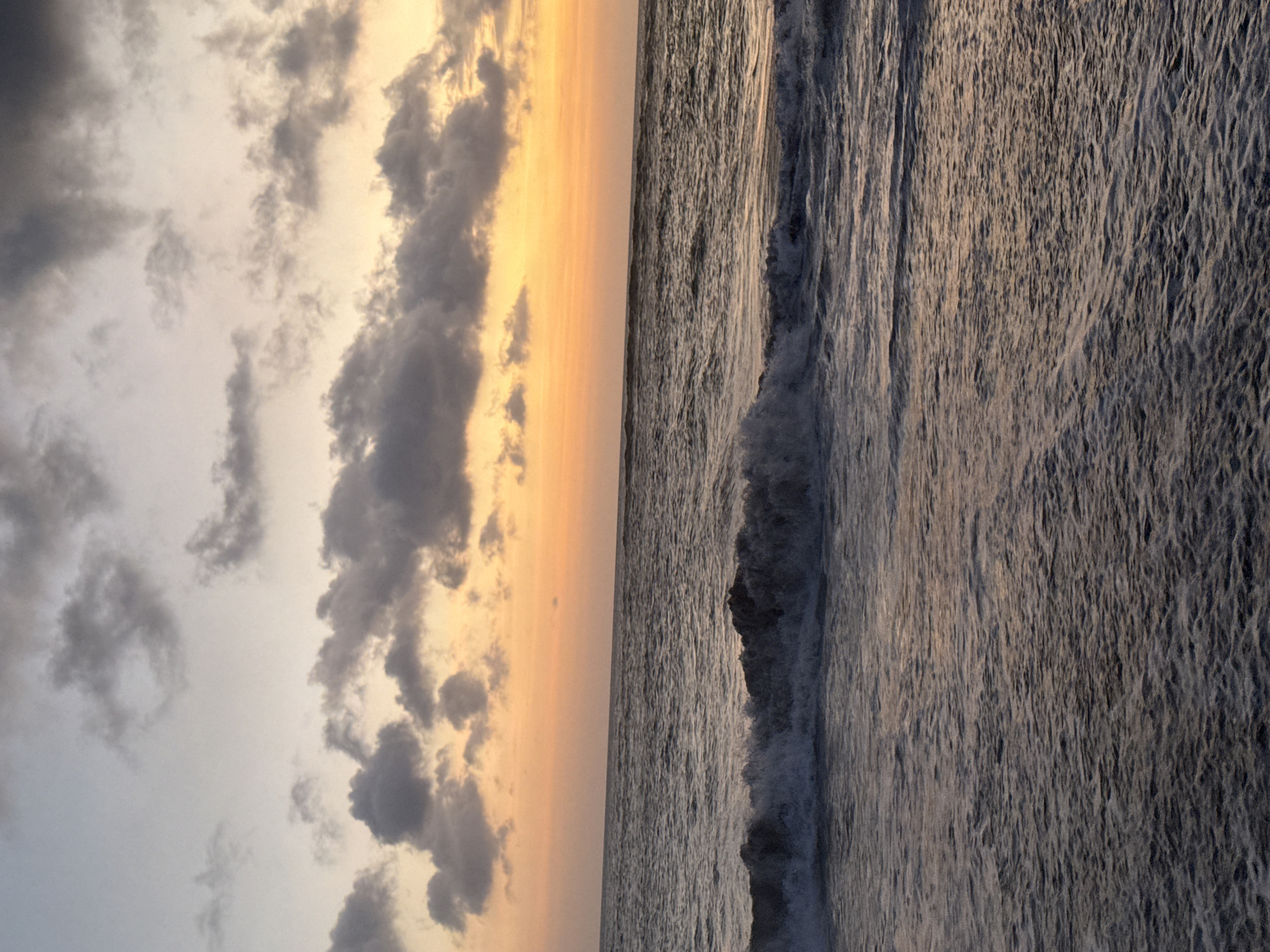 The image shows a beach scene at sunset, likely along a coastline, in an unknown city and state. The sky is filled with a combination of dark gray clouds and golden-orange light from the setting sun. The clouds are denser towards the left side of the image and gradually fade to a lighter tone as they approach the center. The ocean water is visible on the right side of the image. Waves are present, with the closest wave showing the breaking of water. The water appears dark gray in color.
