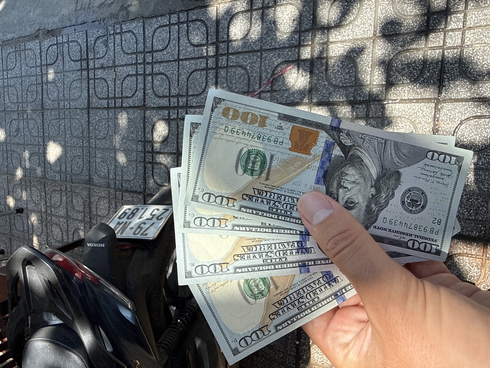 In Nha Trang, Vietnam, a person's hand holds a fan of five US hundred-dollar bills, obscuring a portion of a tiled sidewalk and the rear of a dark-colored motorcycle. The motorcycle's license plate reads "79-NA 251.89". The bills are slightly worn and exhibit creases, with the serial number "PB93874439Q" visible on one. The scene is illuminated by natural sunlight, casting dappled shadows on the ground. The overall context suggests a transaction or display of money in a public urban setting.