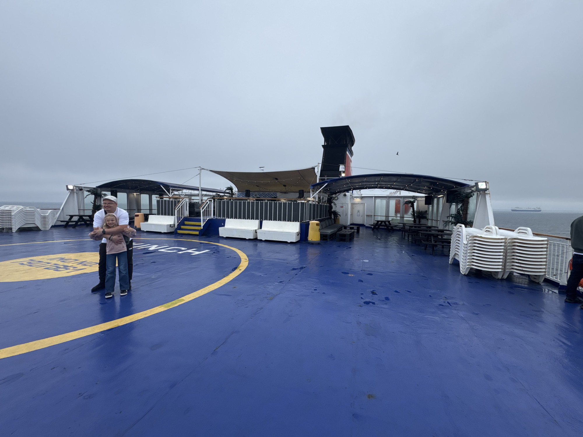 The image depicts a man and a young girl standing on the blue deck of a ferry. The man, wearing a white cap and a dark jacket, has his arms around the girl, who is in a pink hoodie and blue jeans. They are positioned within a yellow circular marking on the deck, which appears to be part of a helipad.
In the background, there are covered seating areas with tables and chairs, as well as stacks of white lounge chairs. A large smokestack with red and black markings rises from the ferry. The sky is overcast, and the sea is visible in the distance with a large cargo ship on the horizon. The ferry appears to be docked or at sea under a cloudy sky.
The scene does not provide any visual cues to specifically locate it within the city of Gdynia, Poland.