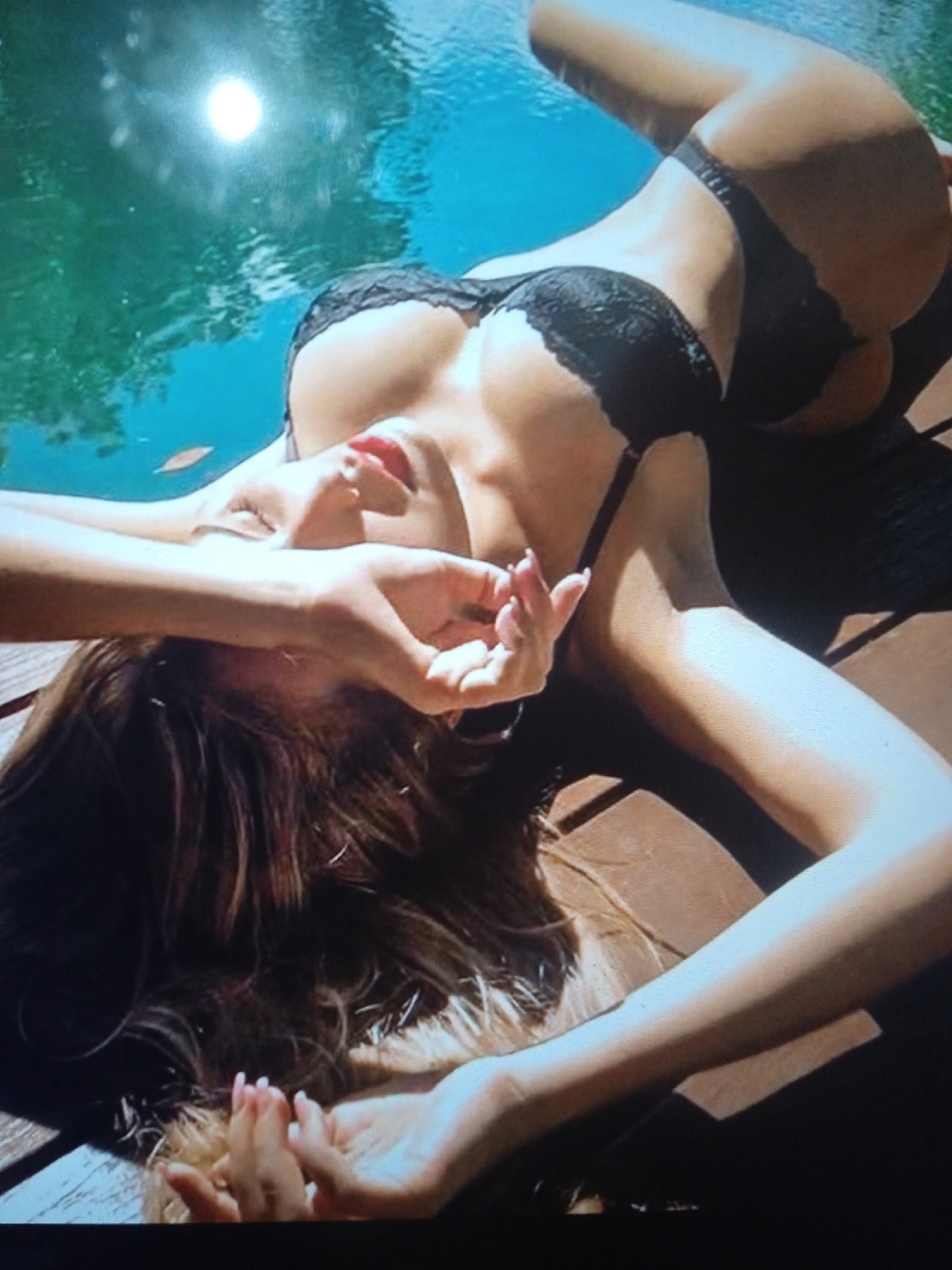 The image shows a person lying on their back on a wooden surface, near a body of water. The person is wearing a black bikini. Their face is partially obscured by their hands, and their eyes are closed. The water appears to be a pool, with sunlight reflecting off the surface. The person's body is positioned in a relaxed manner, with one leg bent and the other extended. The overall scene evokes a sense of leisure and relaxation. There is no information available to suggest this image is located in Chernivtsi, Ukraine.
