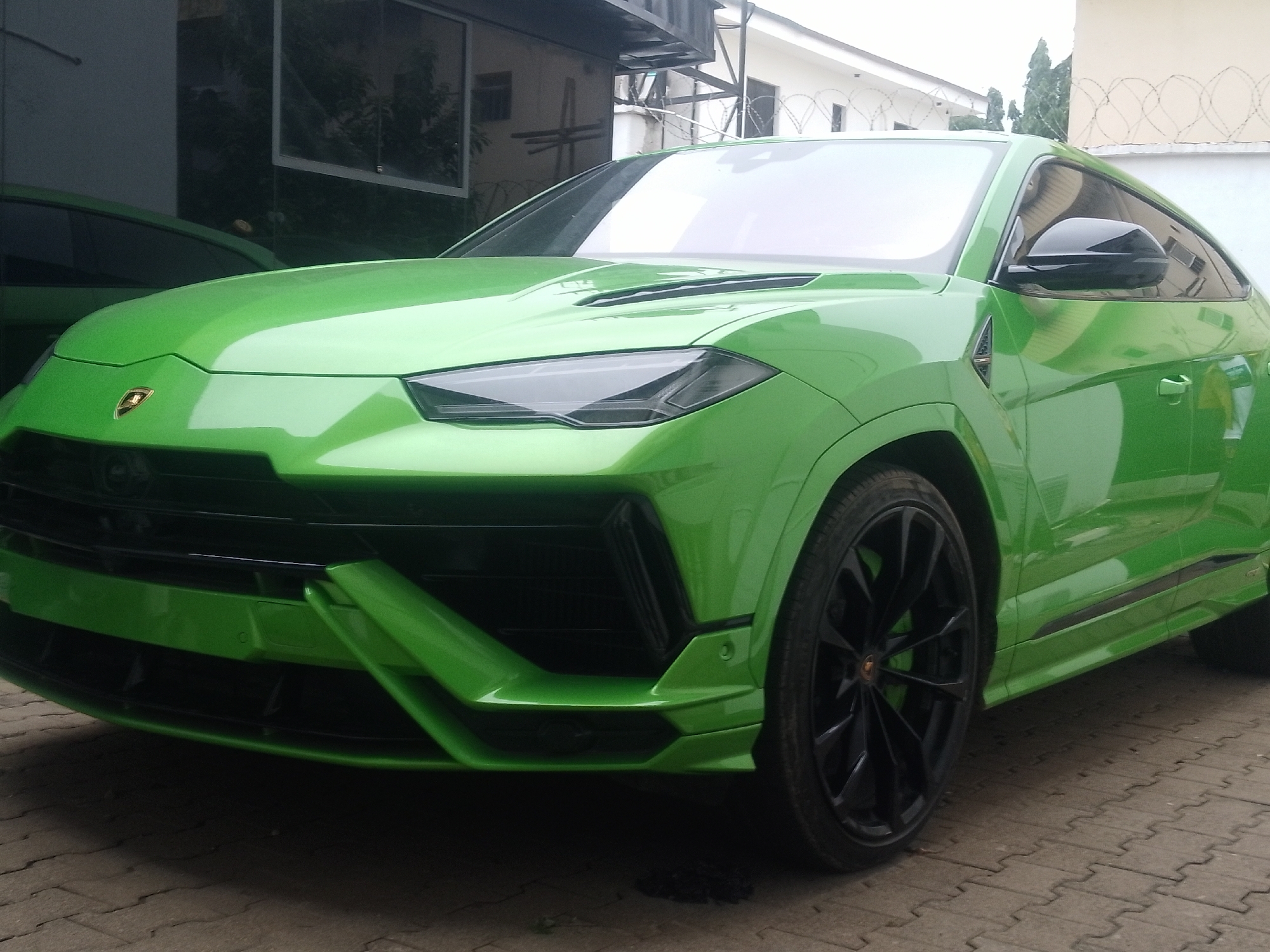 An eye-level, medium shot captures the front left quarter of a bright green Lamborghini Urus parked on a patterned concrete surface. The car is facing forward, with its headlights angled slightly inward, illuminating a dark grille and aggressive front bumper. A prominent Lamborghini emblem is centered on the hood. The vehicle's exterior reflects the surrounding environment, including a building with windows and a metal structure overhead. The background reveals a partially visible second green car to the left and a white building with barbed wire fencing. The scene appears to be an outdoor location in Abuja, Nigeria.