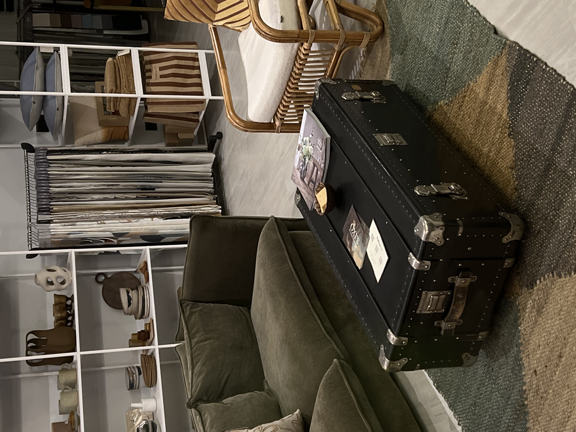 The image depicts an indoor scene, likely a retail environment or a staged living space, showcasing a collection of home decor items. The primary subjects are objects, including a large, black, vintage-style trunk with metal accents, a plush sofa in a muted green color, and a rattan chair with striped cushions. Various shelves are filled with decorative items such as pottery, bowls, and stacked textiles. A rug with a geometric pattern in shades of green, brown, and gray is visible on the floor, partially covered by the sofa and trunk. Two small books or magazines are placed on top of the trunk.
There is no discernible activity or event taking place; the scene appears to be static, emphasizing the arrangement of the items. The lighting suggests an indoor setting, possibly during the day or with artificial lighting. The overall mood is one of curated display and interior design.
Notable details include the intricate metalwork on the trunk and the textured weave of the rug. The shelves hold a variety of items, suggesting a focus on artisanal or handcrafted goods. The style of the furniture and decor hints at a contemporary or eclectic aesthetic.
Visible text on the books or magazines on the trunk includes "OXIS" and some smaller, less legible text. The location context provided is Sofia, Bulgaria.