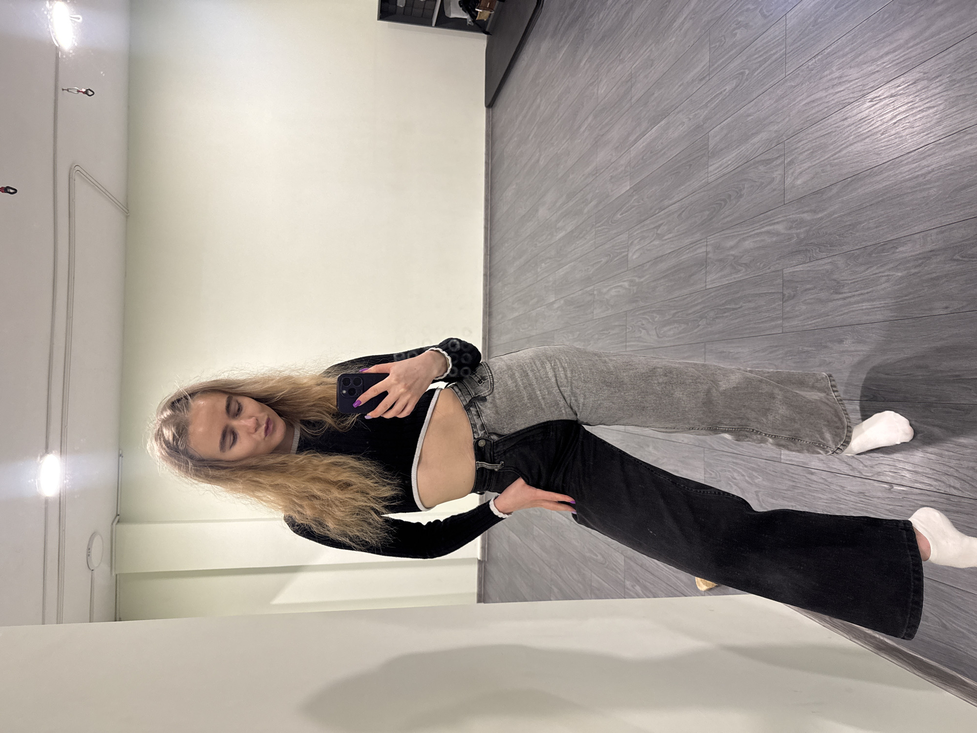 A young woman with long blonde hair is taking a selfie in a room. She is wearing a black long-sleeve crop top with a white trim and mismatched jeans, with one leg grey and the other black. She is holding a dark-colored smartphone in her left hand, her face turned slightly towards the camera. Her fingernails are painted purple. The room has a white wall and a gray wood-like floor. There are light fixtures on the wall and storage above her. The location is Unknown, Unknown.