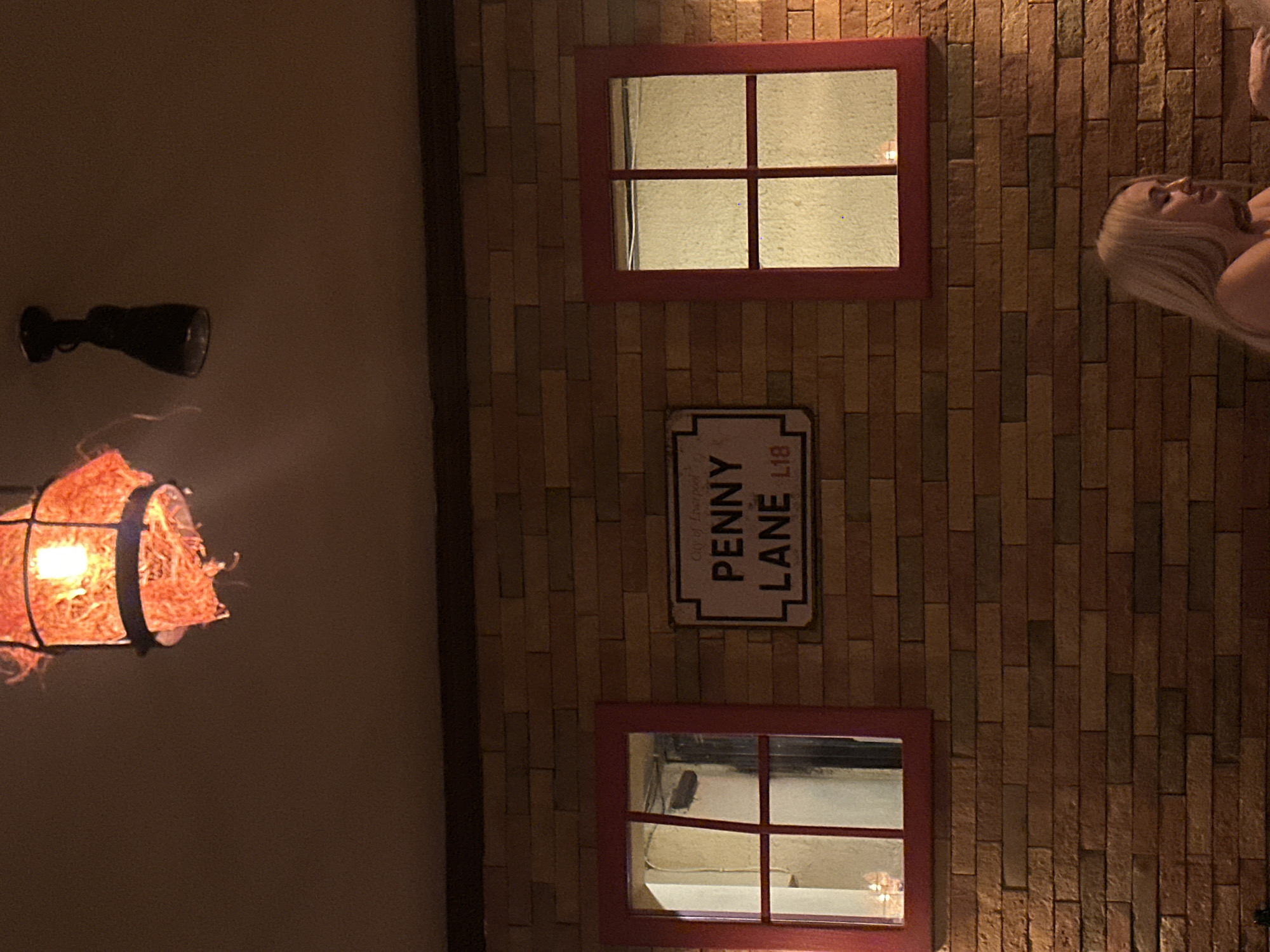 The image depicts an interior setting with a wall resembling a brick facade. Two red-framed windows with divided panes are positioned above and below a "Penny Lane L18" street sign. To the left, a decorative lamp with a straw-like covering illuminates the scene. A person with blonde hair is partially visible on the right side of the frame. The scene does not appear to be in Yerevan, Armenia, but rather suggests a stylized interior setting, possibly related to The Beatles or the Penny Lane area of Liverpool, England.